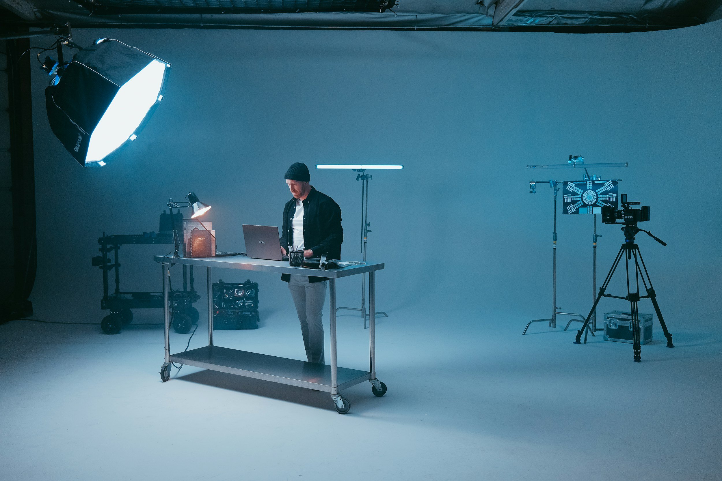 A man wearing a black beanie and black jacket working on a laptop at a table in a photo studio with professional lighting equipment and a light blue backdrop.