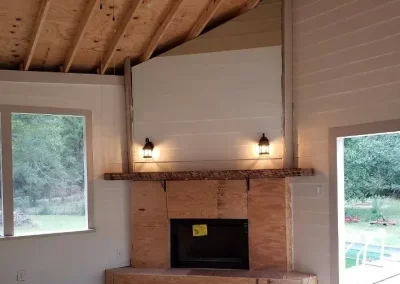 Living room with a vaulted ceiling, brick fireplace, and two wall-mounted lamps above the mantel. Large windows on the left and right sides show outdoor scenery.