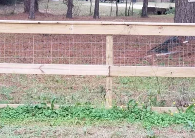 A wooden fence with wire mesh in front of a grassy yard, trees, and a house in the background.