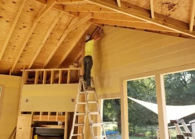 People working on interior construction of a house with exposed wooden framing and a ladder.