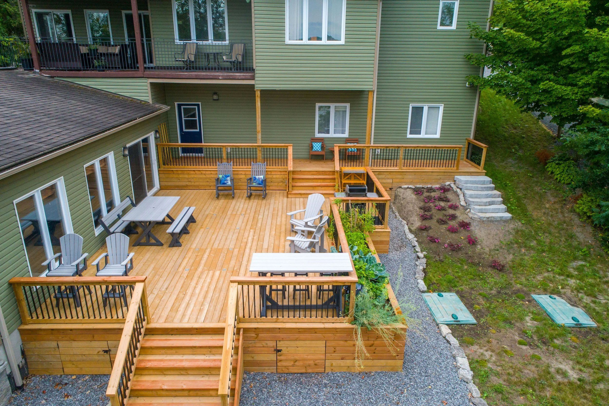 A multi-level wooden deck attached to a green house with sliding glass doors, outdoor chairs, tables, and a grill, overlooking a landscaped backyard with a flower bed, stairs, and a tree.