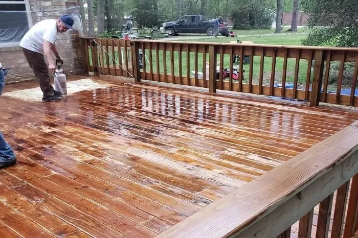 A person is sealing a new wooden deck with a waterproofing product while on a rainy day. The deck is outside a house, with a wooden railing, and a backyard with some trees and a parked truck.