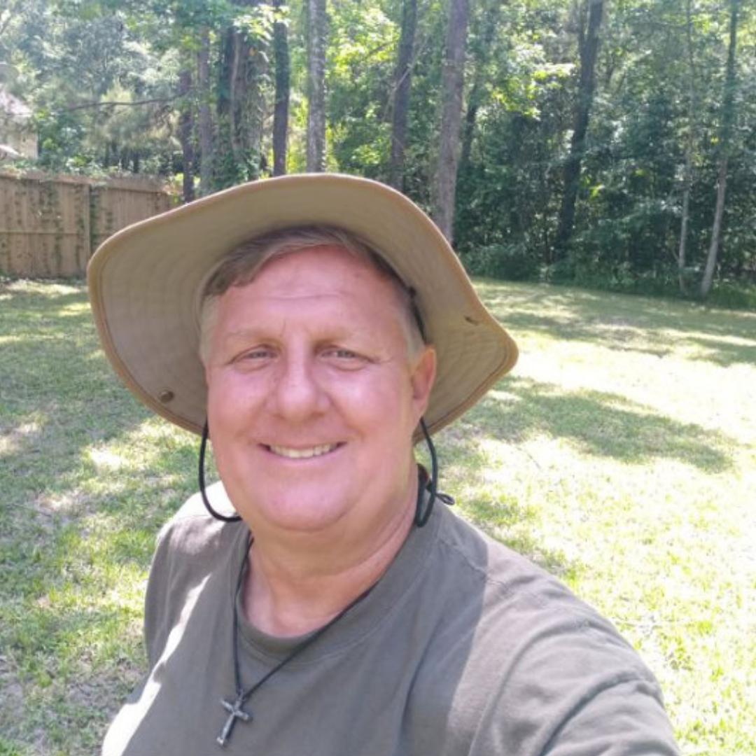 A smiling man outdoors in a grassy backyard with trees, wearing a wide-brimmed hat, olive green shirt, and a cross necklace.