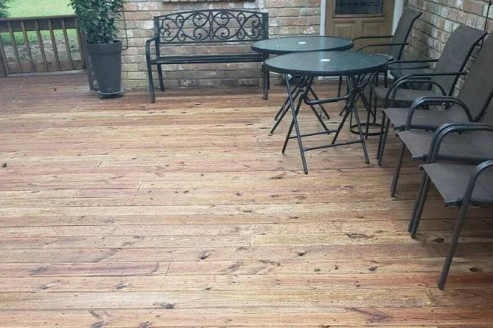 Outdoor wooden deck with a black metal bench, two round black tables, four black chairs, a potted plant, and a brick wall in the background.