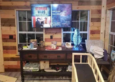 A wooden interior with a large flat-screen TV displaying Star Wars images, a window, and a table with decorations and snacks. There are shelves underneath with various items, and stacked blankets or towels on a chair.