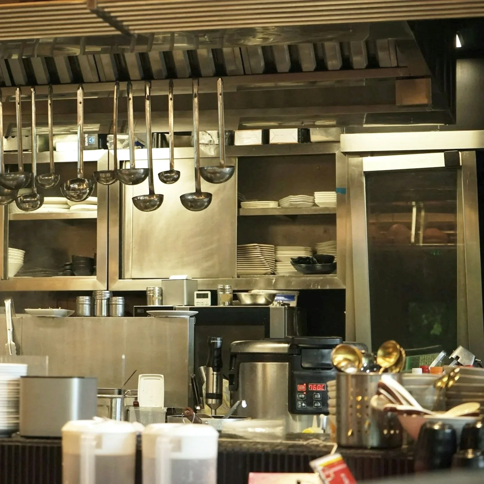 A chef wearing a white uniform, black face mask, and tall white hat working in a commercial stainless steel kitchen with various pots, pans, and kitchen equipment.