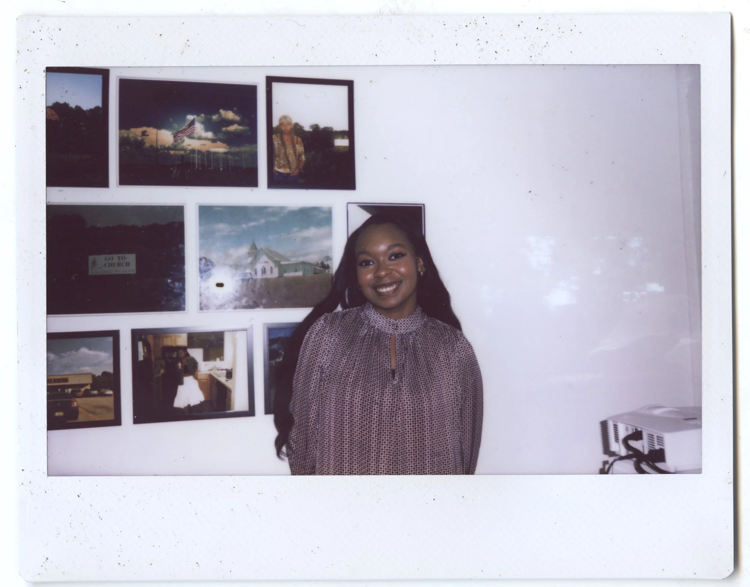 A Polaroid photo featuring a smiling young woman with dark hair, standing in front of a wall decorated with various photographs and images.