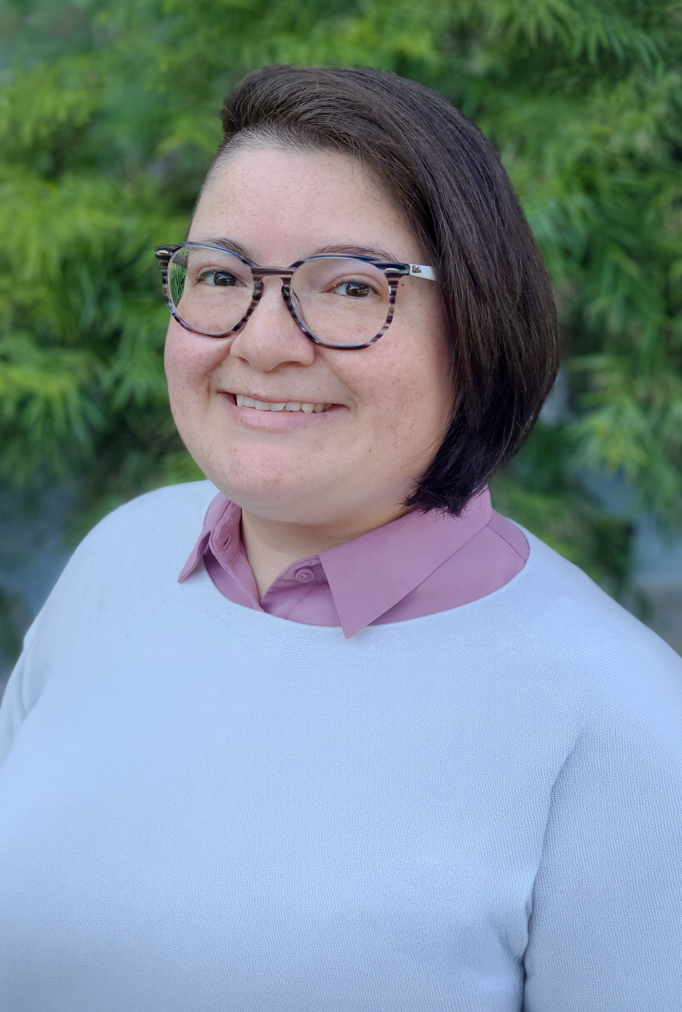 Close-up portrait of a smiling woman with short dark hair, wearing glasses and a peppermint pink collared shirt layered under a white sweater, standing outdoors with blurred green foliage in the background.