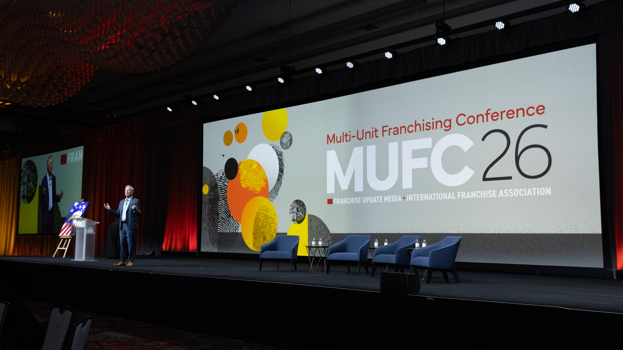 A man in a suit standing on a stage in front of a large screen displaying the words 'Multi-Unit Franchising Conference MUFC 26' and the subtitle 'Franchise Update Media International Franchise Association'. There are unoccupied chairs and a small table on stage, with a portrait on an easel to the side.