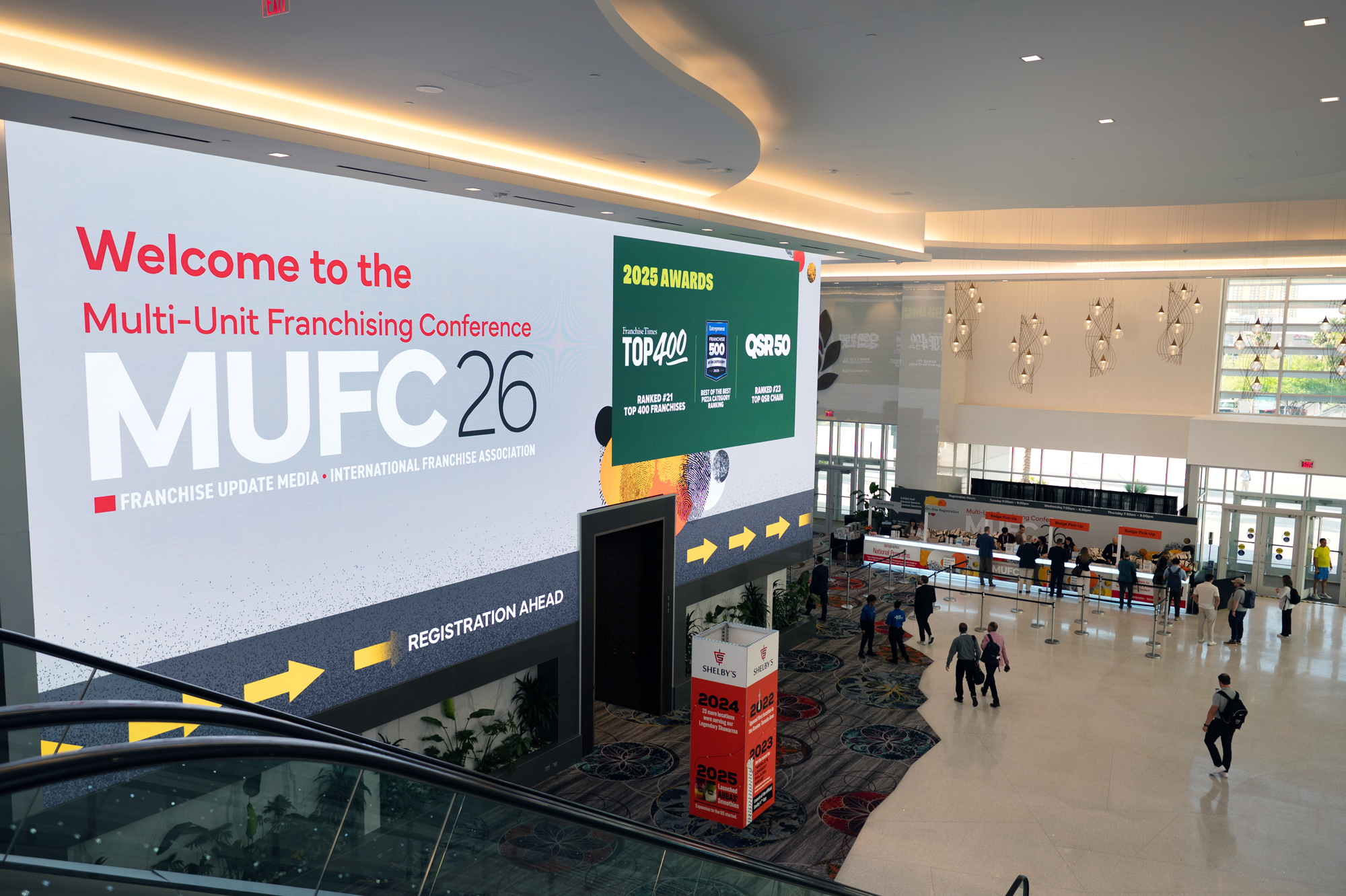 Large digital sign welcoming attendees to the Multi-Unit Franchising Conference (MUFC) 2026, displaying rankings and awards from franchise organizations, with a crowded lobby area below and people walking and queuing for registration.