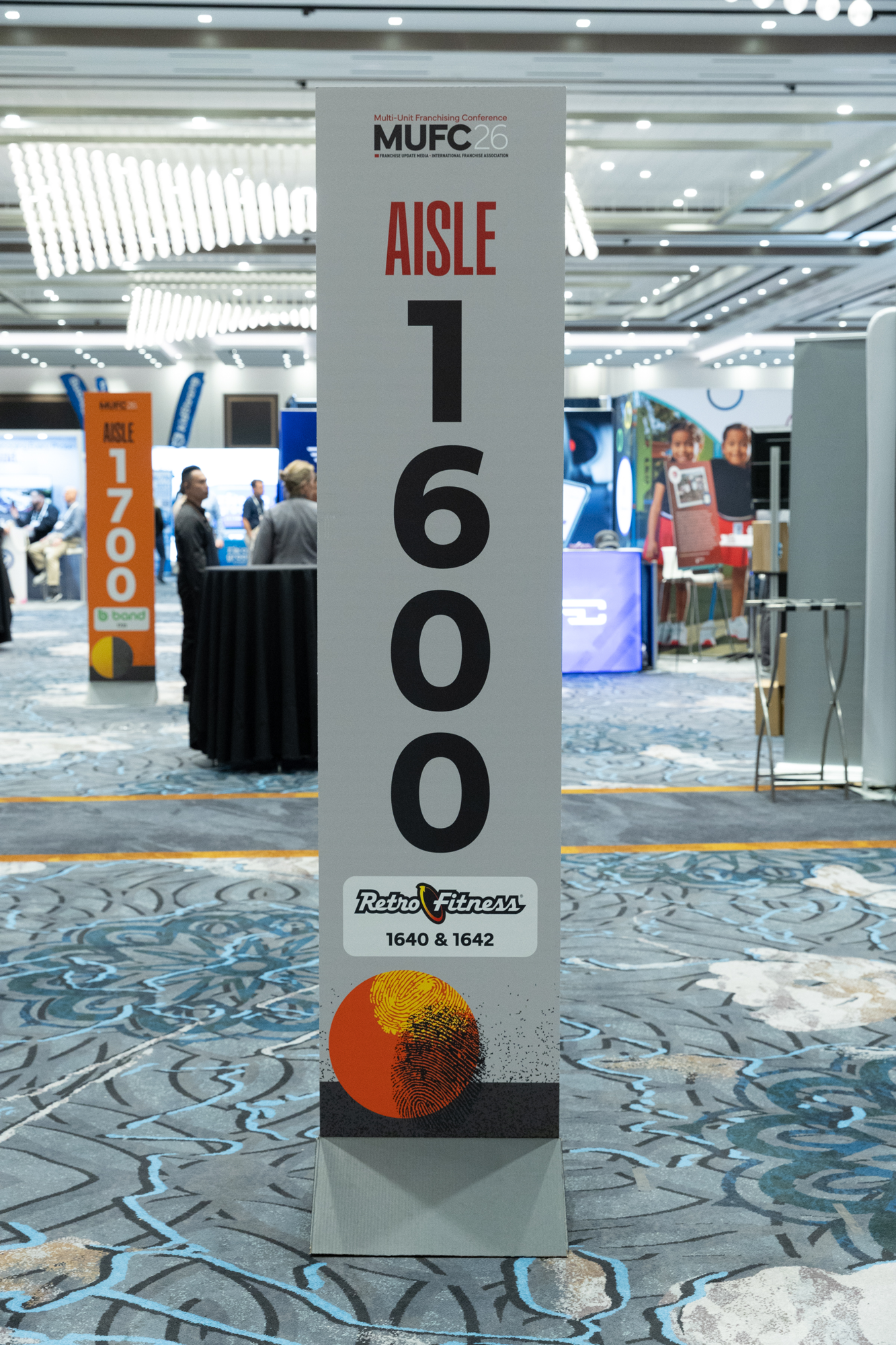 Sign at a conference displaying aisle 1600 for Retro Fitness, with a colorful fingerprint and sun graphic at the bottom. In the background, there are multiple stands and people walking around.