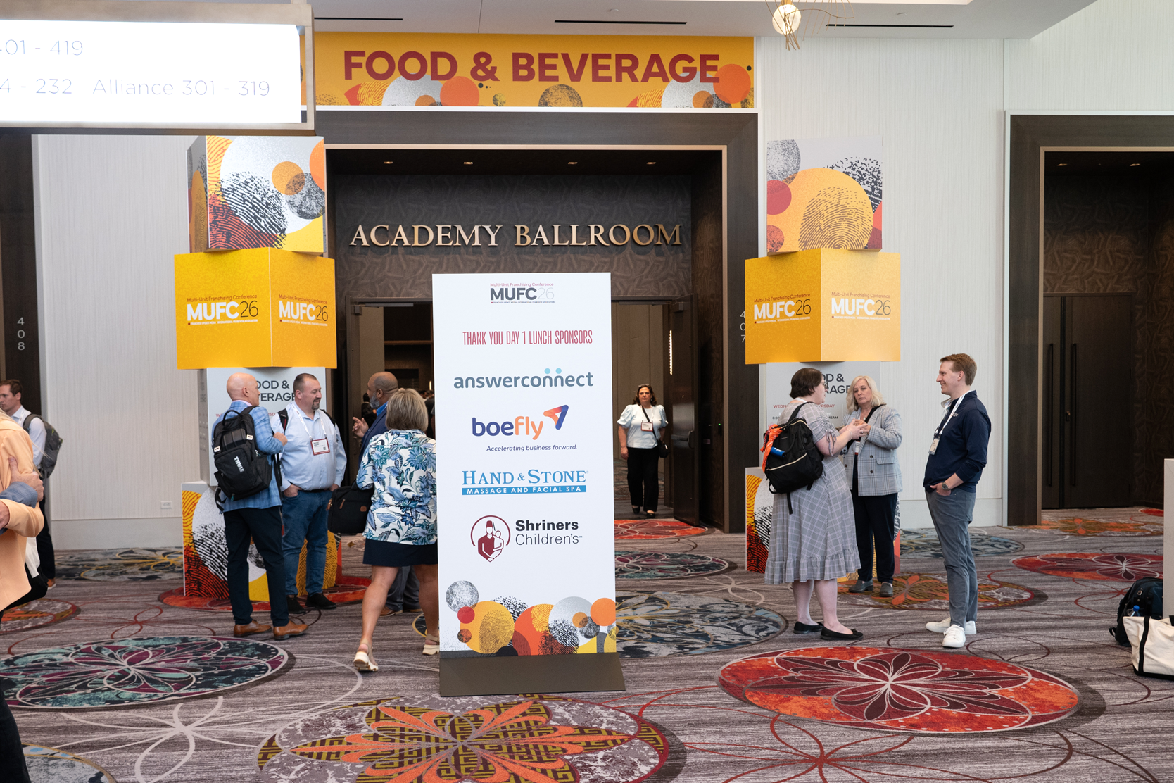 People are gathered outside the entrance of the Academy Ballroom at a conference. There is a tall white sign with sponsor logos and a large white banner inside the doorway thanking lunch sponsors. The area has colorful patterned carpet and decorative wall art with a 'Food & Beverage' sign above.