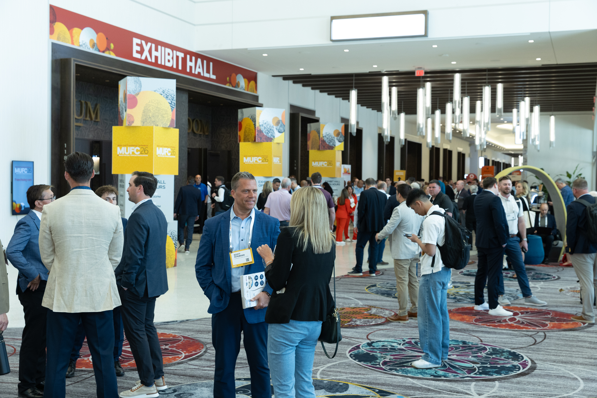 Many people in business attire gathered in a conference hall lobby near an exhibit area, with signs indicating MUFC26 and exhibit hall.
