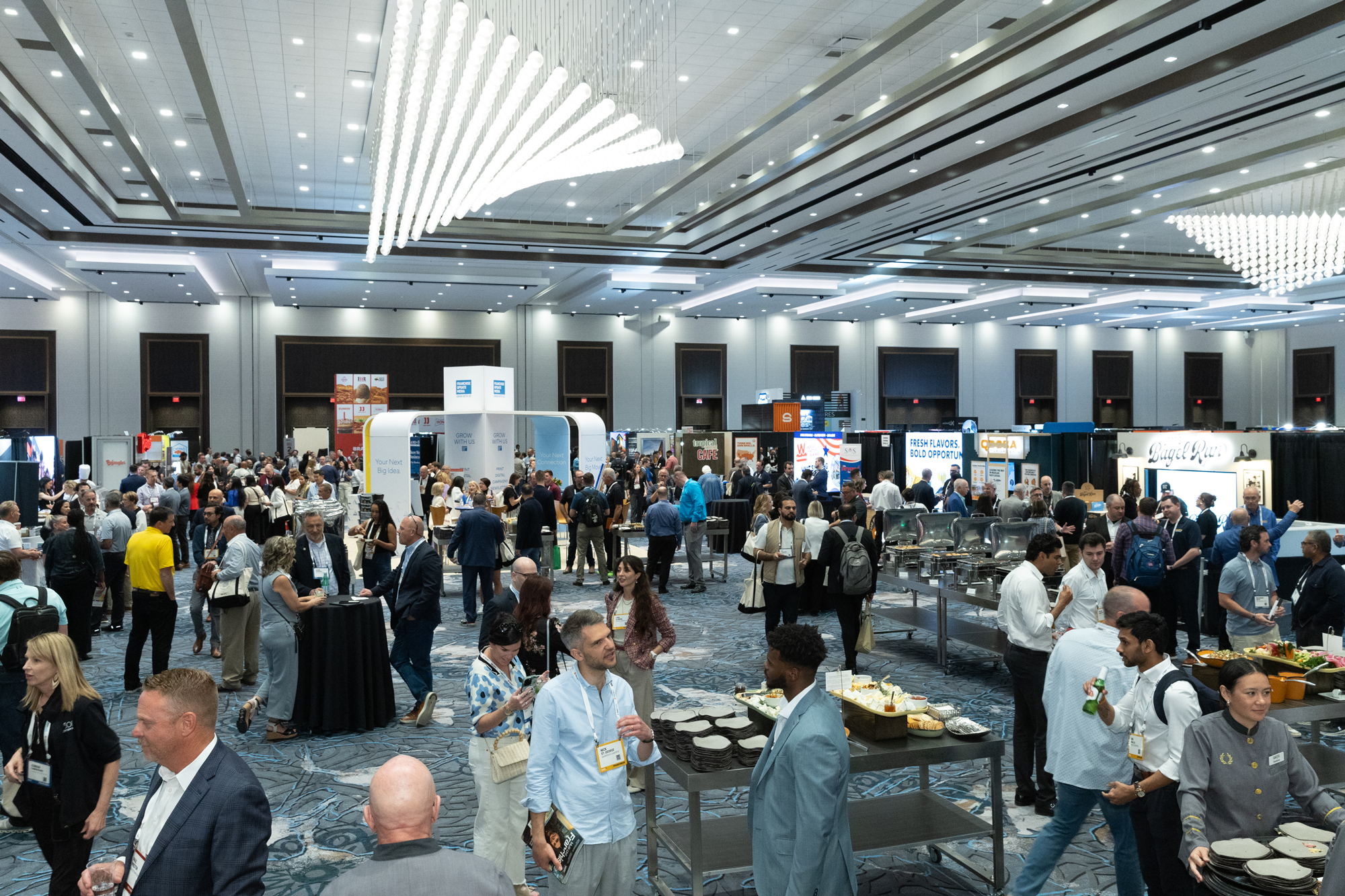 Large conference hall filled with people walking, talking, and standing near tables with food and drinks at a busy business event or conference.
