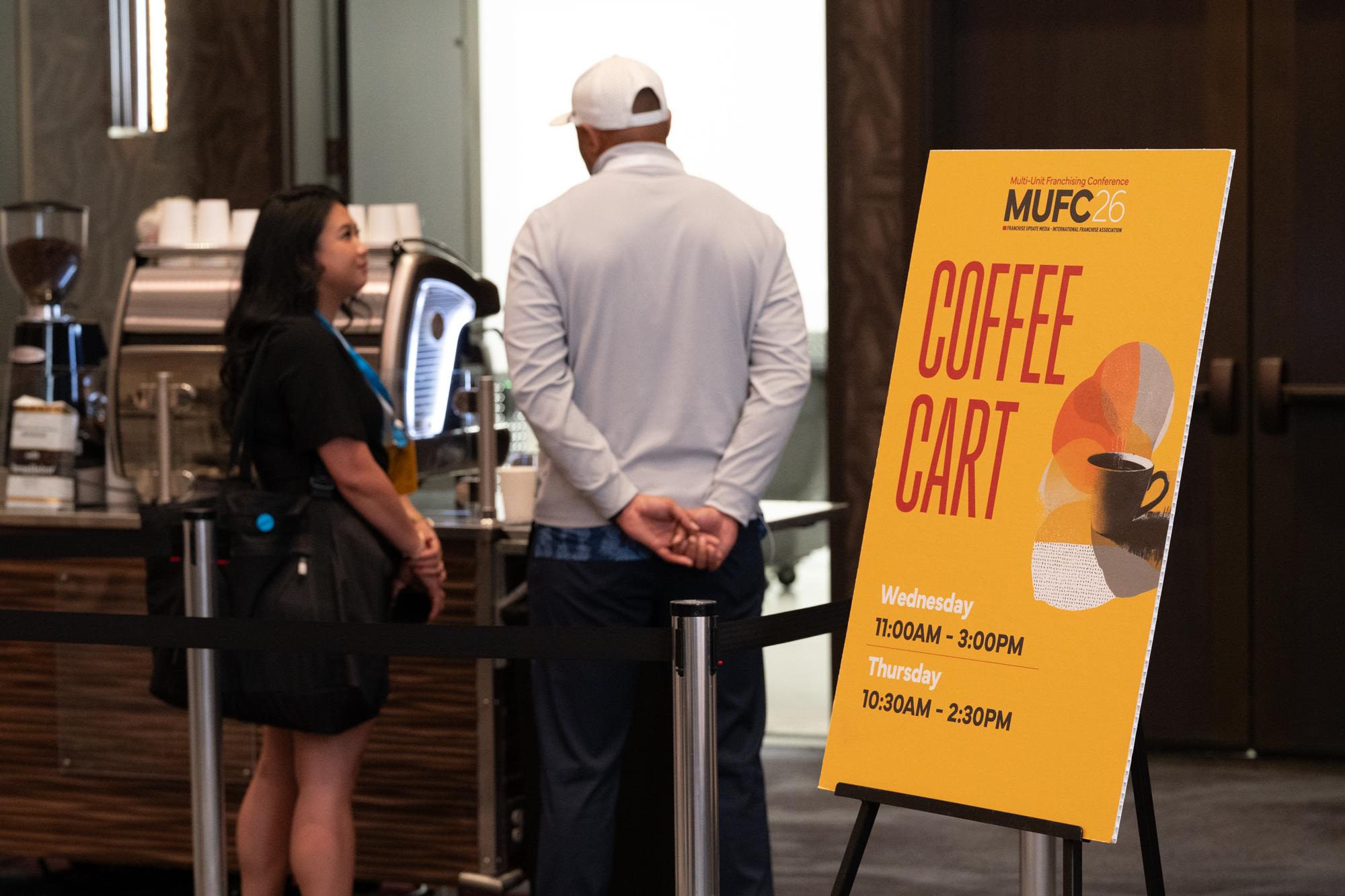 A woman and a man are standing at a café counter, engaged in conversation. The woman is wearing a black dress with a blue lanyard, and the man is wearing a white long-sleeve shirt and a white cap. A yellow sign with information about the coffee cart schedule is in the foreground.