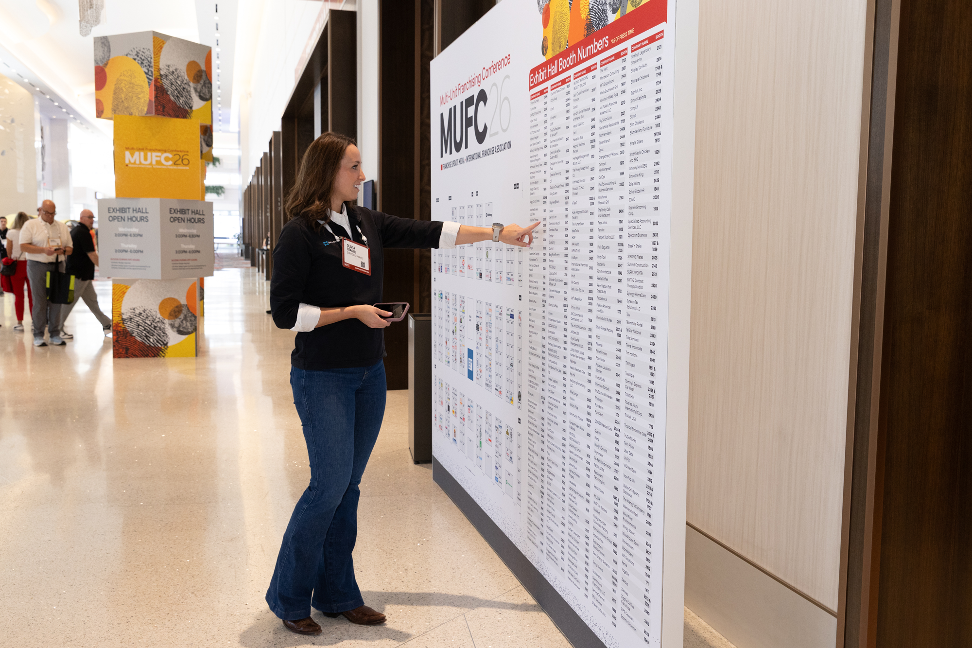 A woman is pointing at a large conference hall booth list on a wall at a business event. She is holding a phone and wearing a name badge. Other people are walking in the background near exhibit signage.