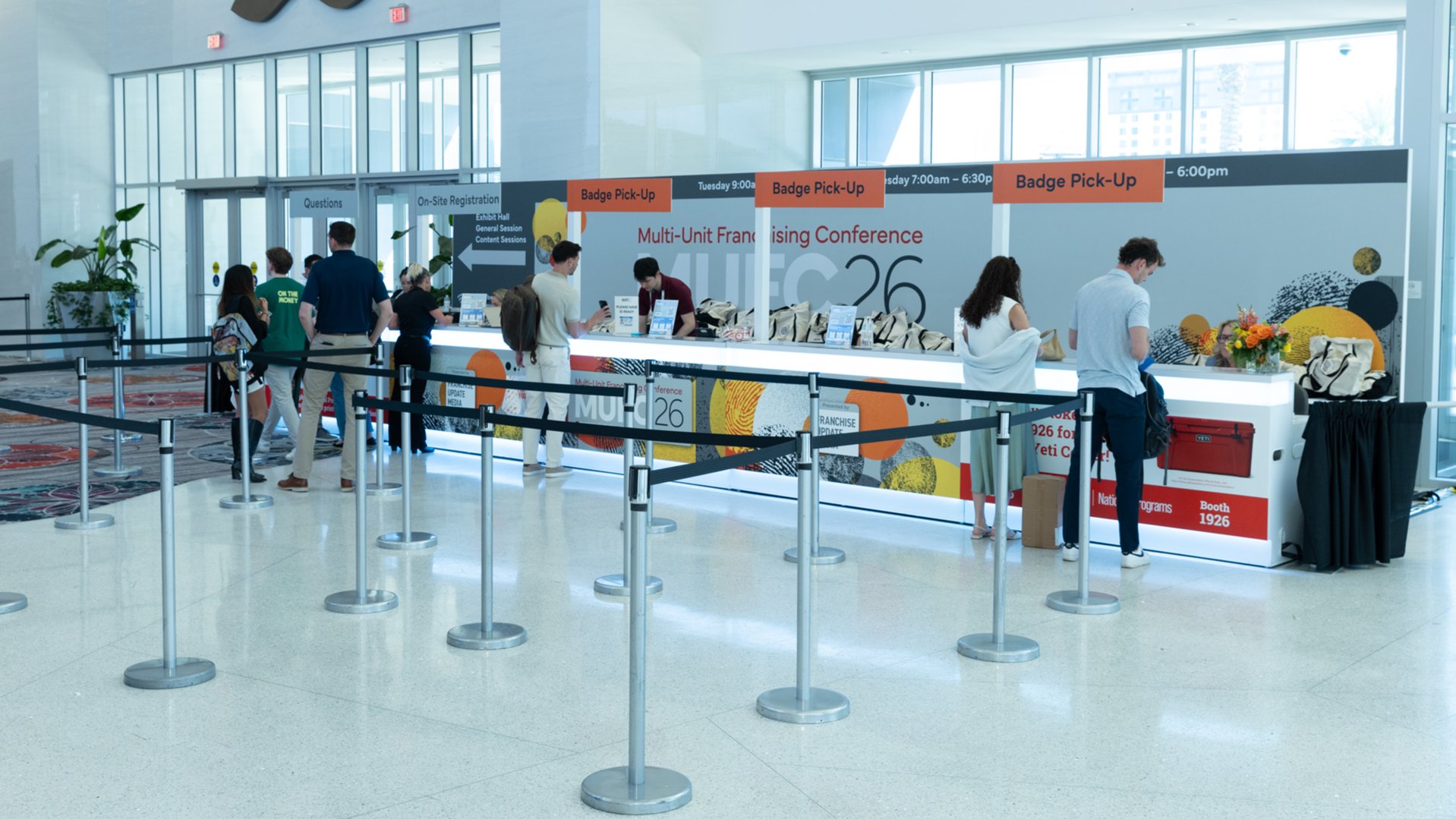 Registration area at a conference, with people standing in line and at the reception desk, bright lighting, large windows, floral arrangement, and informational signs overhead.