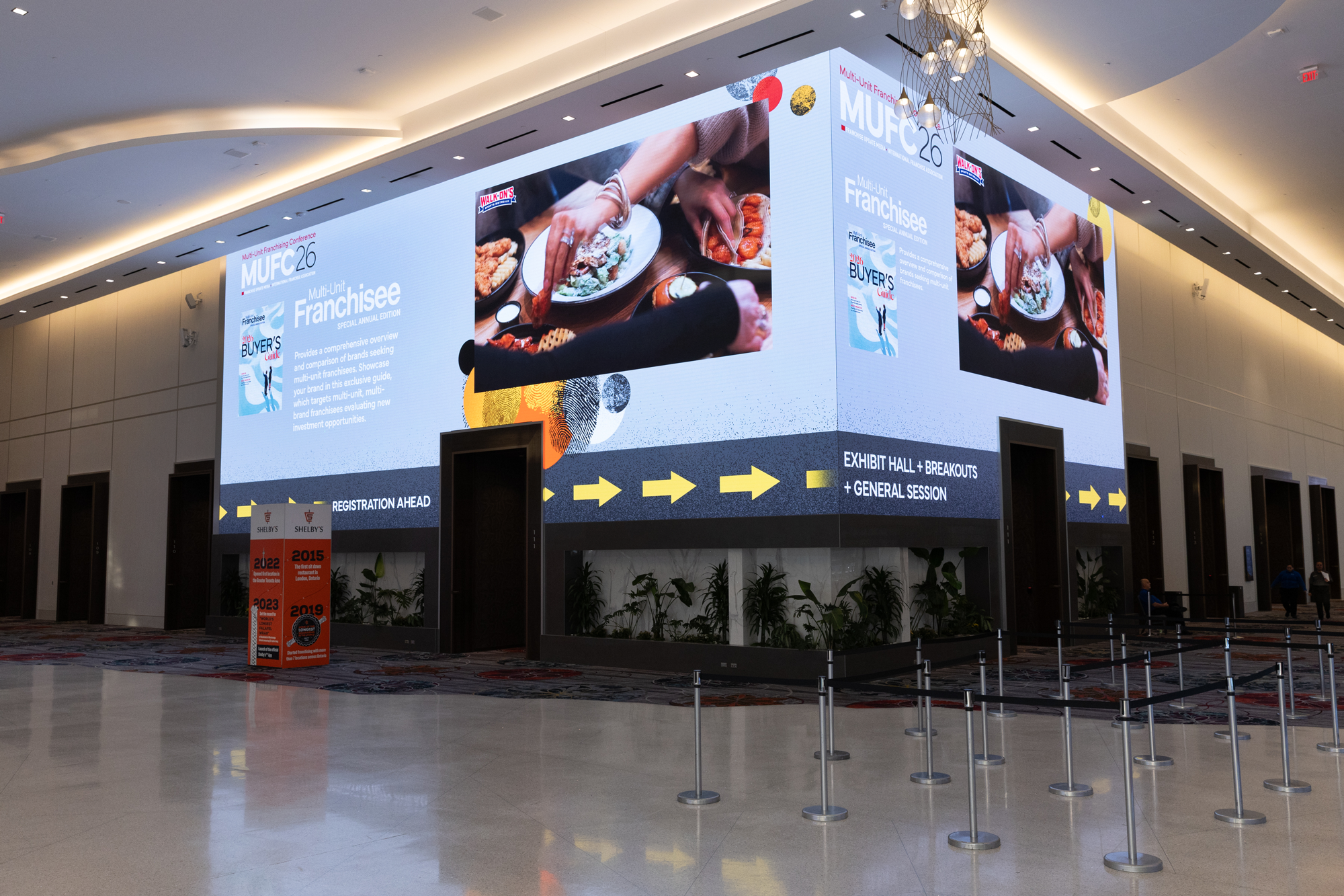 Large digital display at a conference showing event details and food images, with a seating area and a barrier in front.
