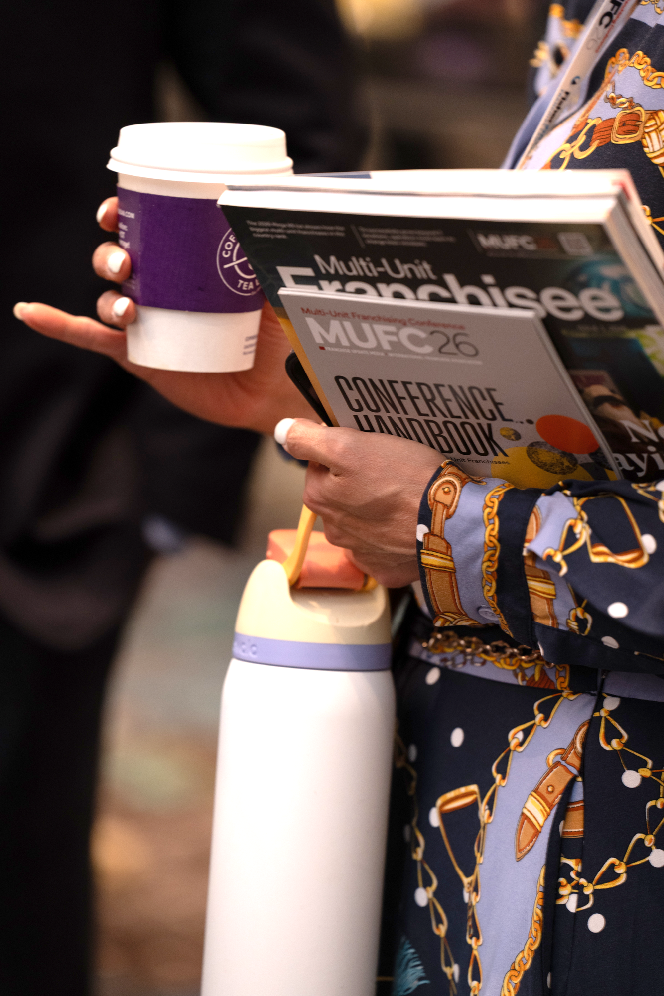 A person holding a paper coffee cup with a purple sleeve and a disposable lid in one hand, and a conference guidebook titled 'Multi-Unit Franchisee Conference Handbook MUFC26' in the other hand. The person is wearing a blue shirt with gold chain and buckle patterns.