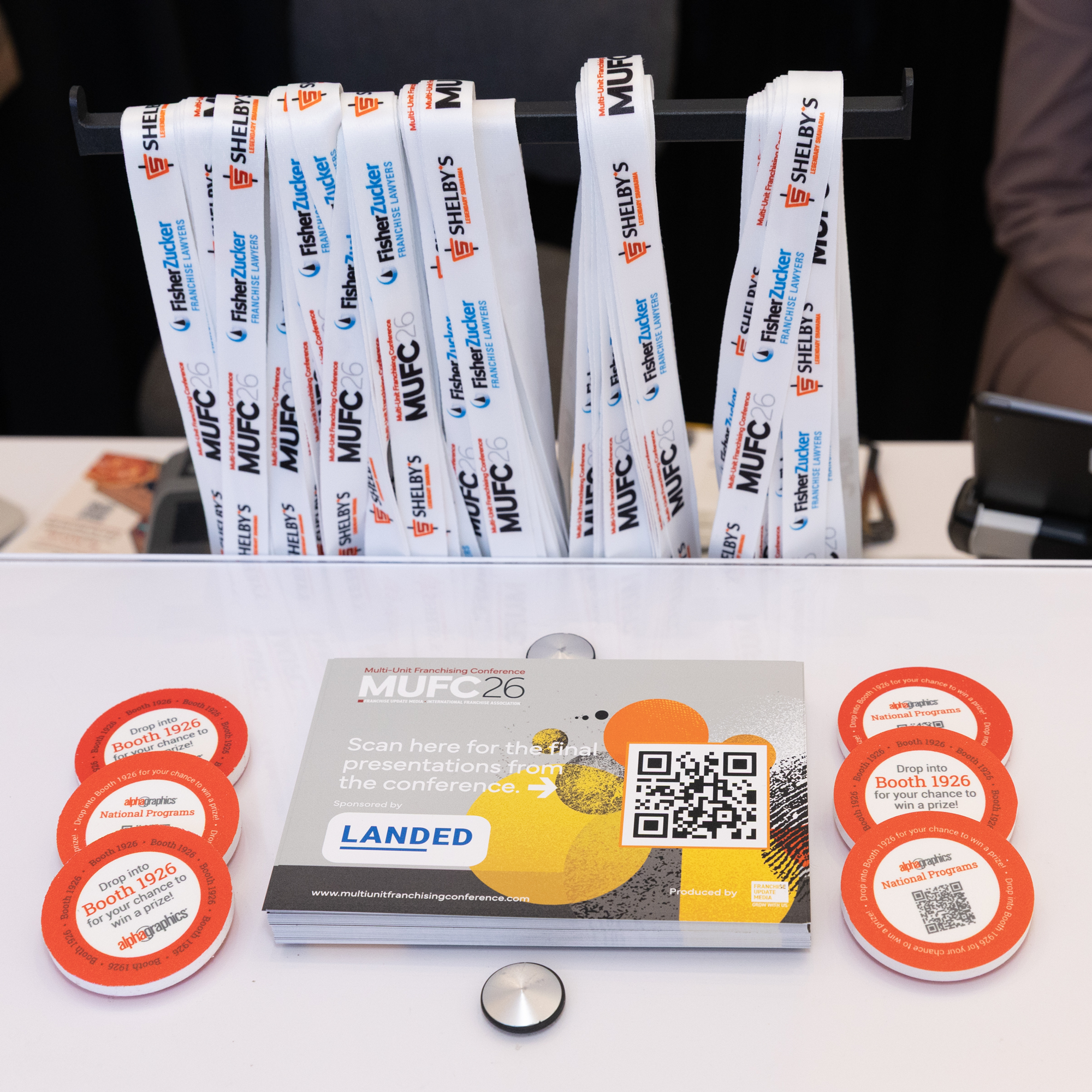 Promotional display of conference materials including lanyards, stickers, and flyers for the Multi-Unit Franchising Conference. The lanyards are hung on a black rack, with the stickers and flyers on a white table with a QR code on one of the flyers.