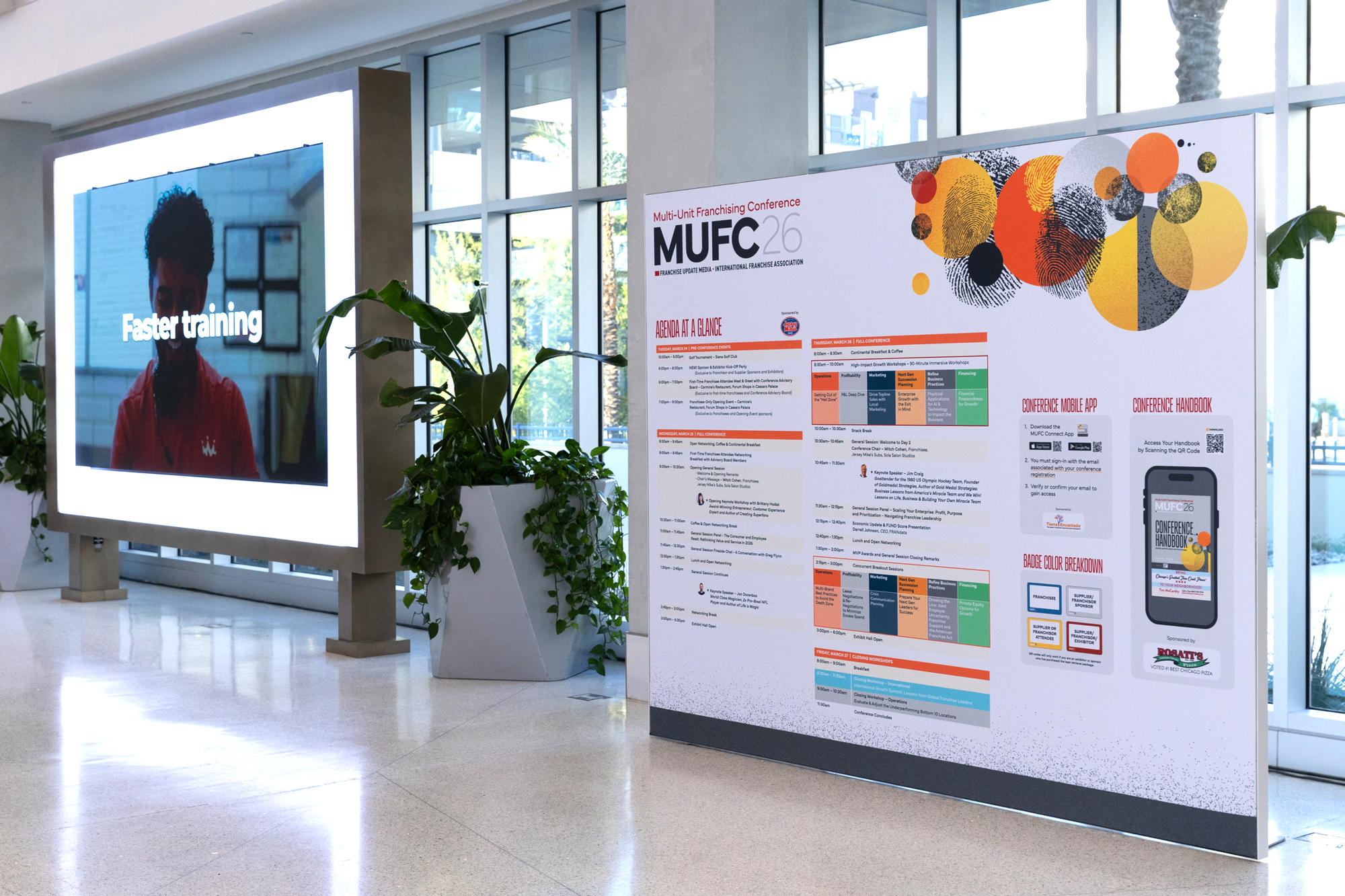 Conference event schedule and signage at a multi-unit franchising conference, with a large digital screen displaying a smiling person's face and text 'Faster training', and a detailed agenda poster in front of large windows.