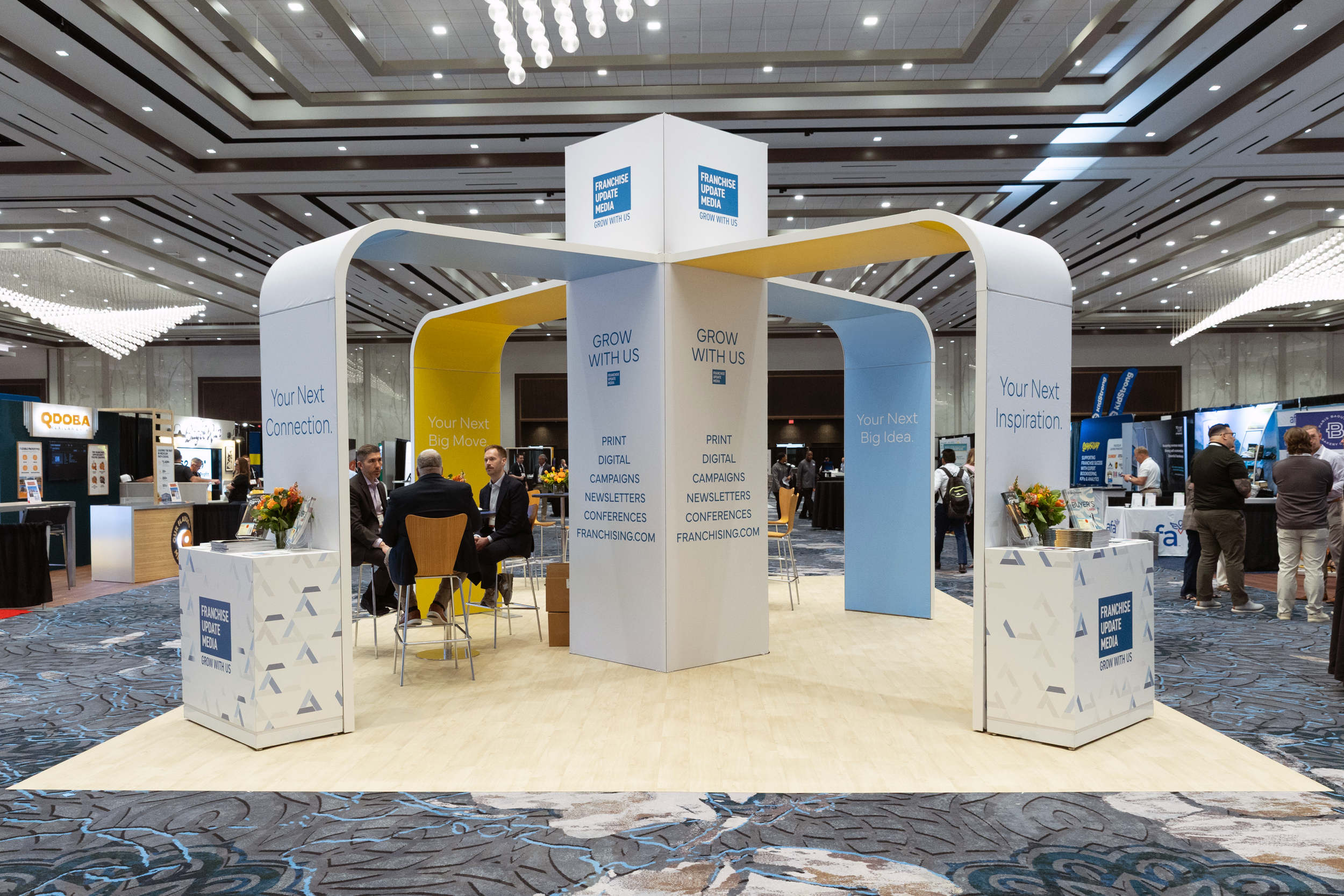 Exhibition booth with signs displaying 'Franchise Update Media' and messages like 'Grow with us', 'Your next connection', 'Your next big move', and 'Your next inspiration'; people are seated and walking around in a large conference hall.