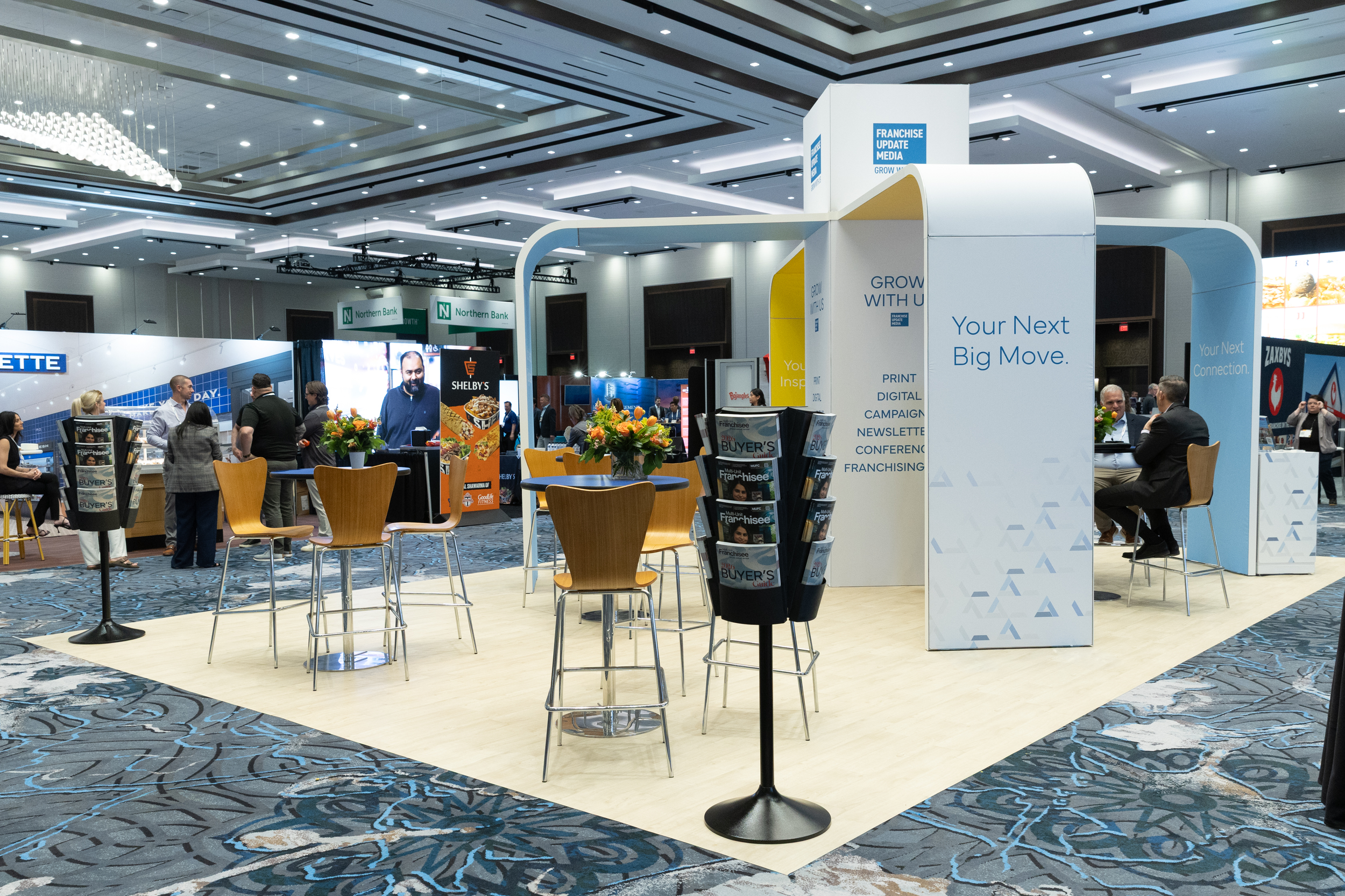 Inside a large conference or exhibition hall with various booths and attendees. Prominent signs include 'Franchise Update Media,' 'Northern Bank,' and 'Shelby’s.' There are tables with chairs and flower arrangements, and attendees are engaging in conversations. The hall has high ceilings with modern lighting.