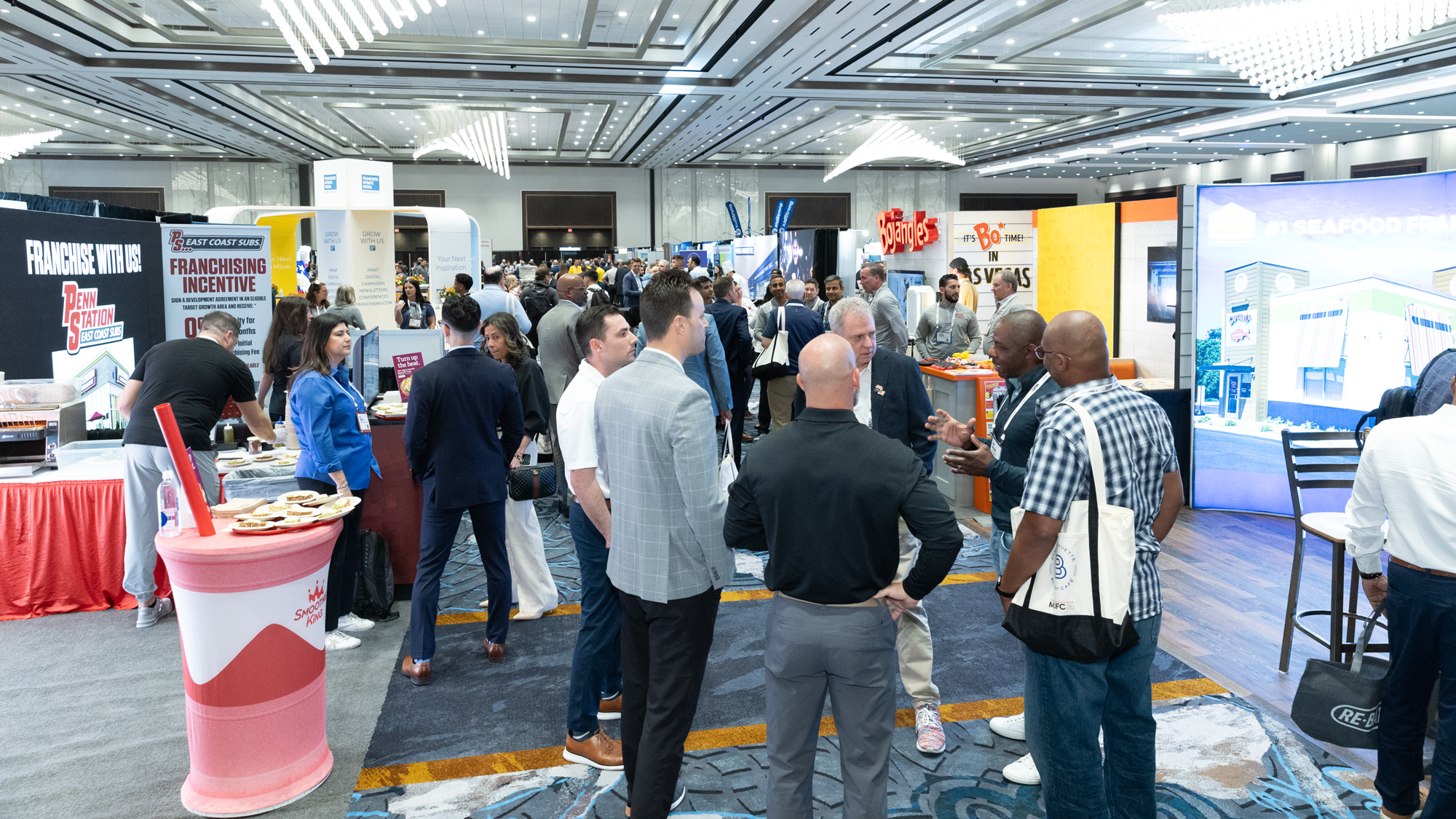 Crowd of people attending a trade show or conference in a large indoor hall with booths and displays.