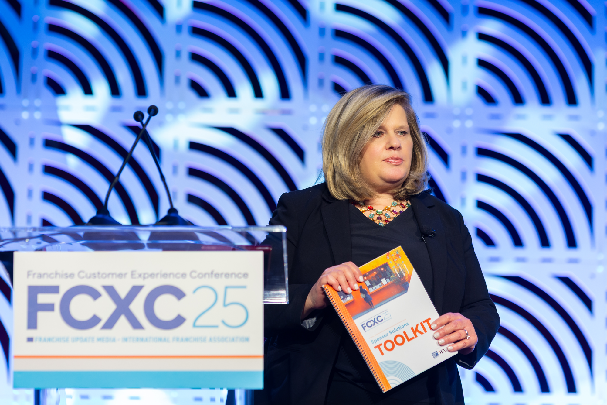 A woman with blonde hair wearing a black blazer and a colorful necklace holding a conference booklet labeled 'TOOLKIT' at a corporate event with blue and purple geometric background.