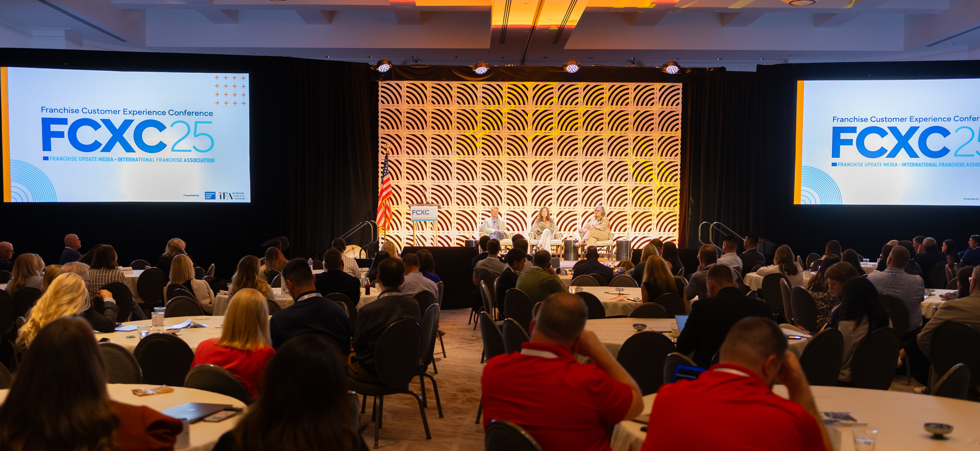 A large conference room filled with attendees listening to a panel discussion on stage. There are two large screens displaying the conference logo 'FCXC 25' and the words 'Franchise Customer Experience Conference'. The stage features three panelists sitting in chairs, with one woman speaking. The room is decorated with black curtains, a patterned backdrop behind the stage, and an American flag. The audience consists of people sitting at round tables, some taking notes or looking at their devices.