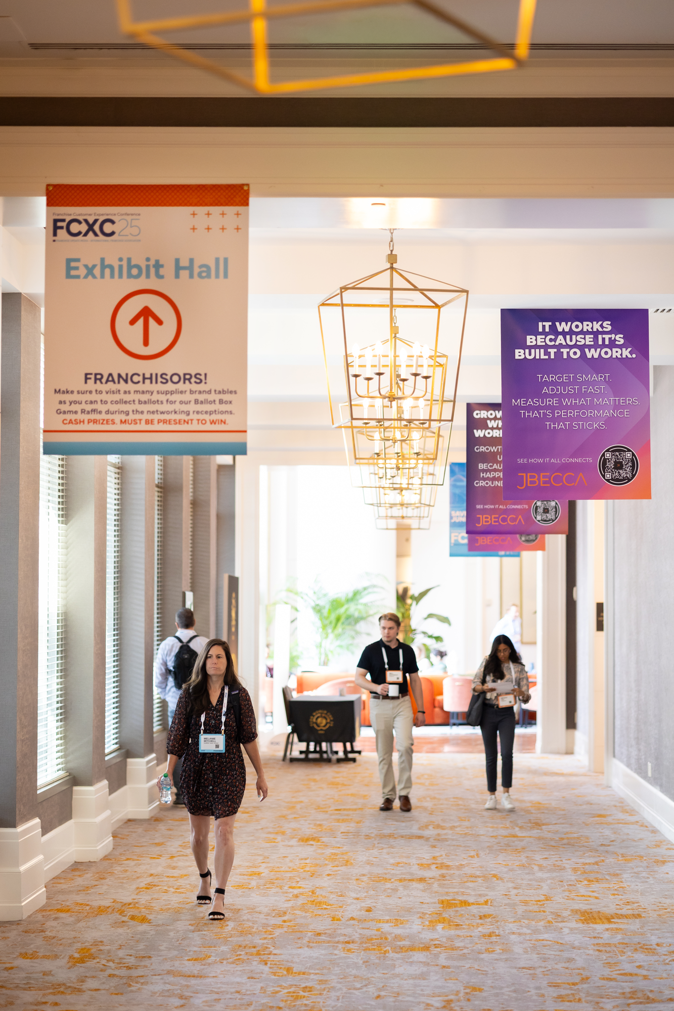 Attendees walking in a conference corridor with banners hanging from the ceiling promoting event activities at the Franchise Customer Experience Conference.