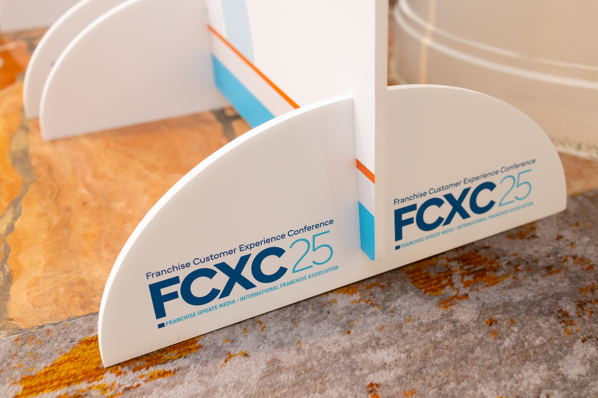 Close-up of a white freestanding display with the logo and text for 'FCXC 25', an international franchise conference, placed on a stone and carpeted floor.