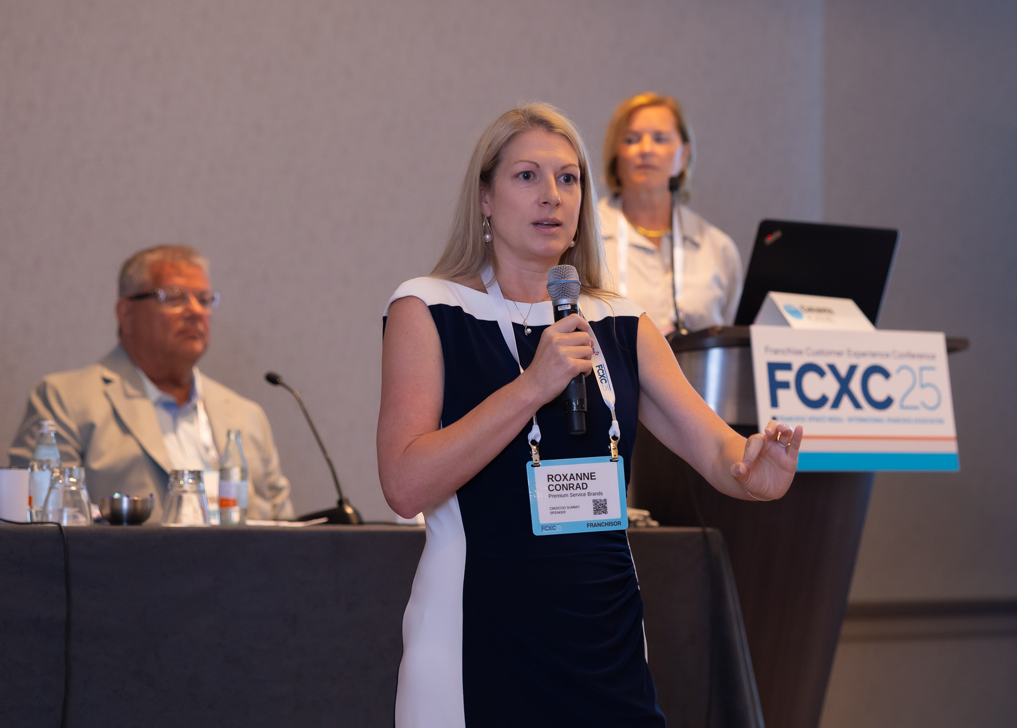 A woman with blonde hair, wearing a black and white dress, holding a microphone, speaking at a conference with a name tag that reads Roxanne Conrad. Behind her, a man in a beige suit and glasses, and another woman standing at a podium with a sign that says FCXC25.
