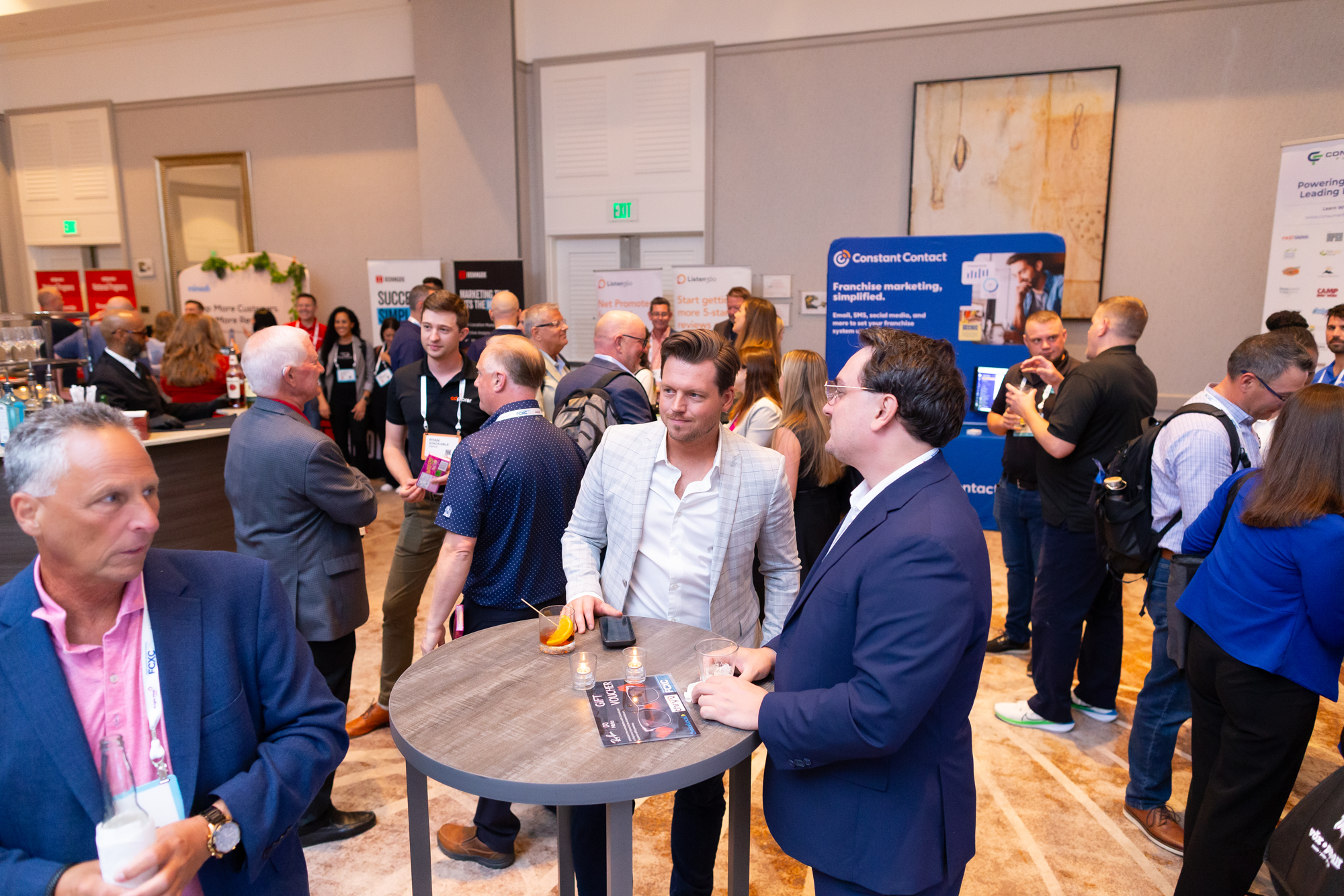 A busy conference scene with people engaging in conversations and browsing booths, including a group gathered around a high-top table with drinks in hand.