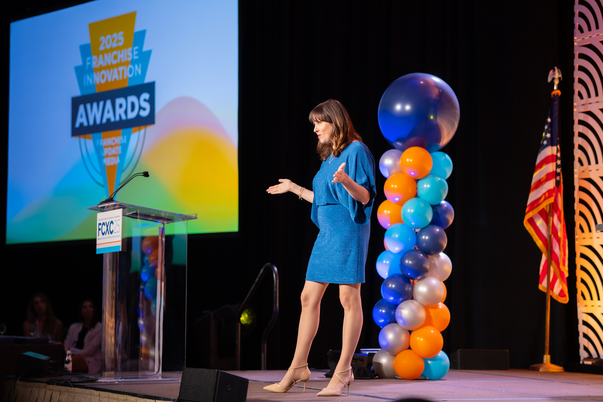 A woman in a blue dress and beige heels on stage during an awards ceremony, with a screen behind her displaying '2025 Franchise Innovation Awards' and colorful balloon decorations nearby.