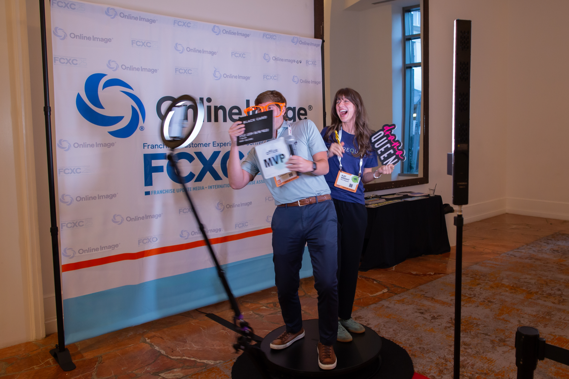 Two people having fun in a photo booth with a FCXC backdrop. The man wears orange glasses, holds a MVP award, and a sign that says 'MVP.' The woman holds a 'Queen' sign and laughs.