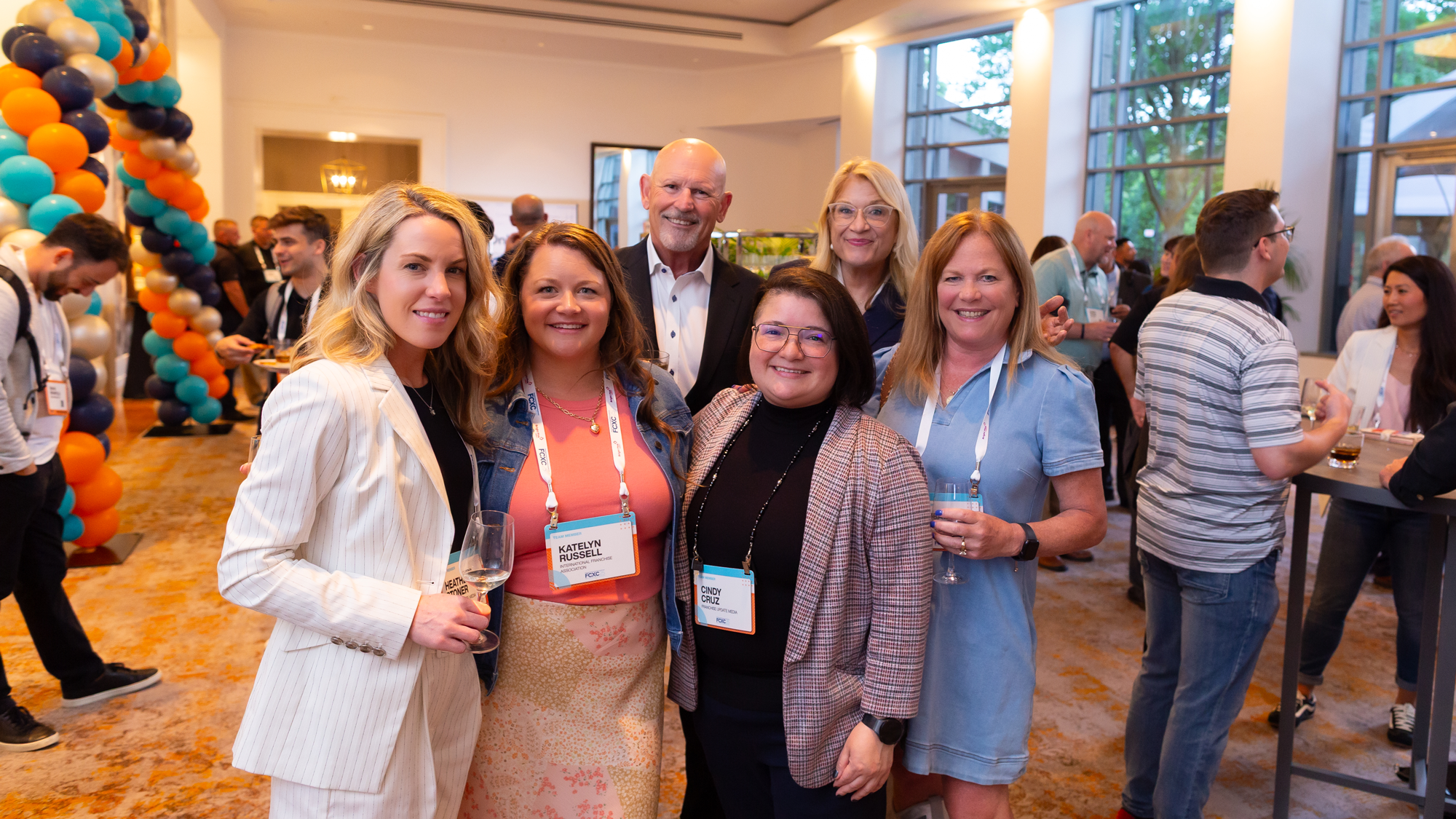 Group of six diverse professionally dressed people smiling at a conference or networking event, standing together in a well-lit venue with large windows and orange, black, teal, and silver balloon decorations.