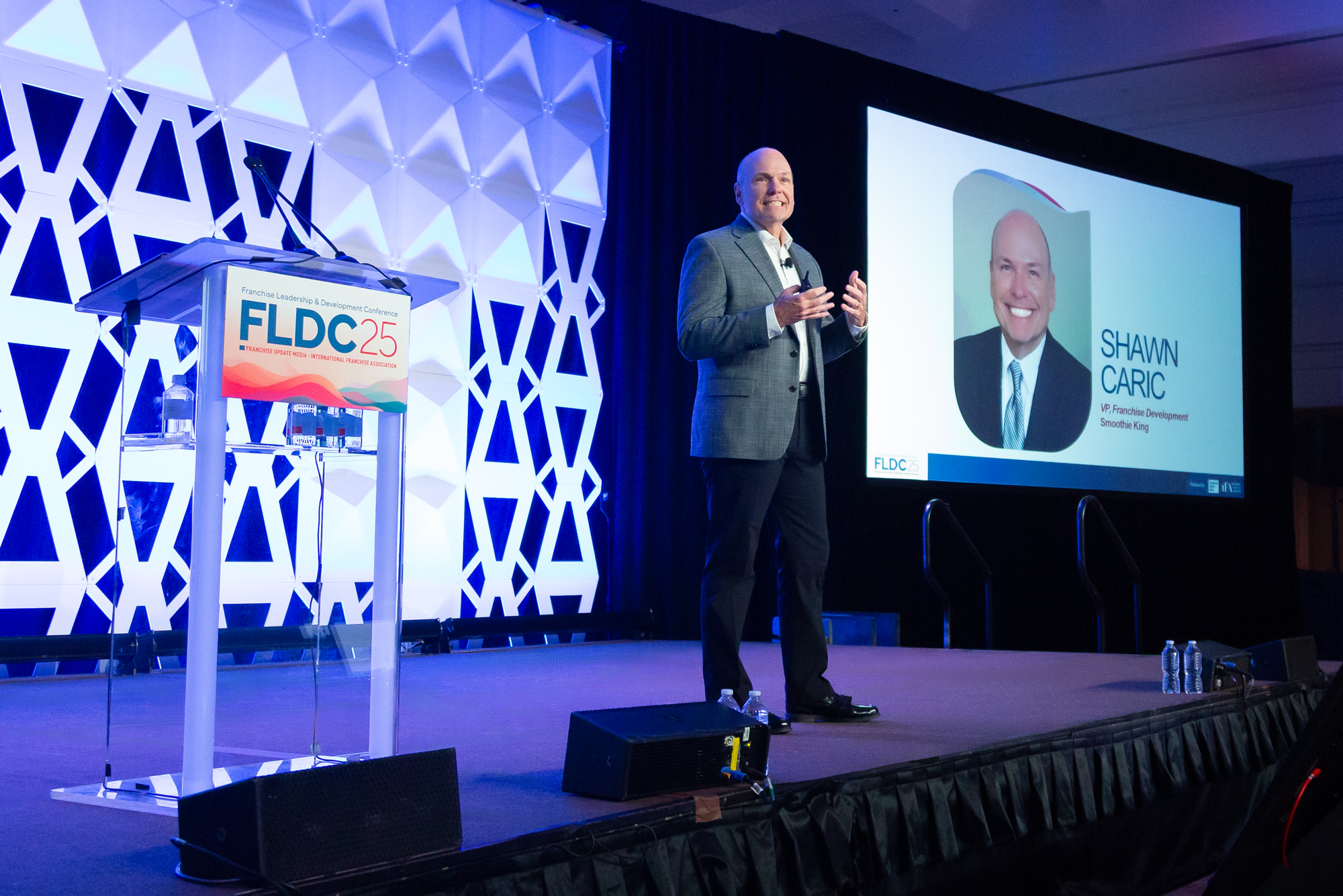 A man in a gray suit presenting on stage at the Franchise Leadership & Development Conference (FLDC) 25. He is standing beside a large screen displaying his photo and name, Shawn Caric, VP of Franchise Development at Smoothie King. The stage has a podium with a sign for FLDC25 and bottles of water, with a patterned white backdrop and dark curtains.
