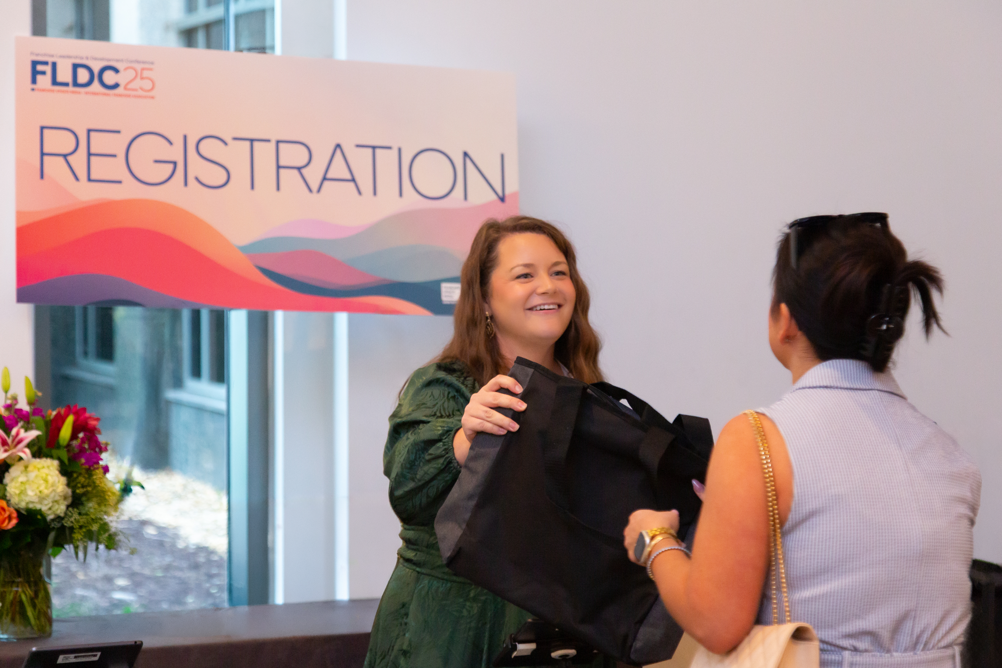 Two women are smiling and greeting each other at a registration desk during an event. There is a sign in the background that reads 'Registration' and a large floral arrangement nearby.
