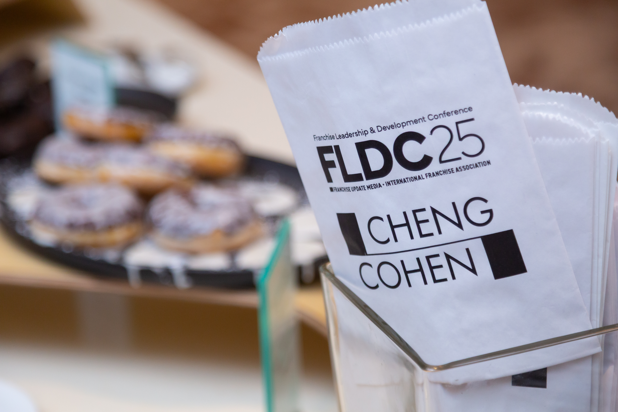 Conference badge with the text 'Franchise Leadership & Development Conference, FLDC25, Cheng, Cohen' on a white lanyard, and a blurred image of donuts on a tray in the background.
