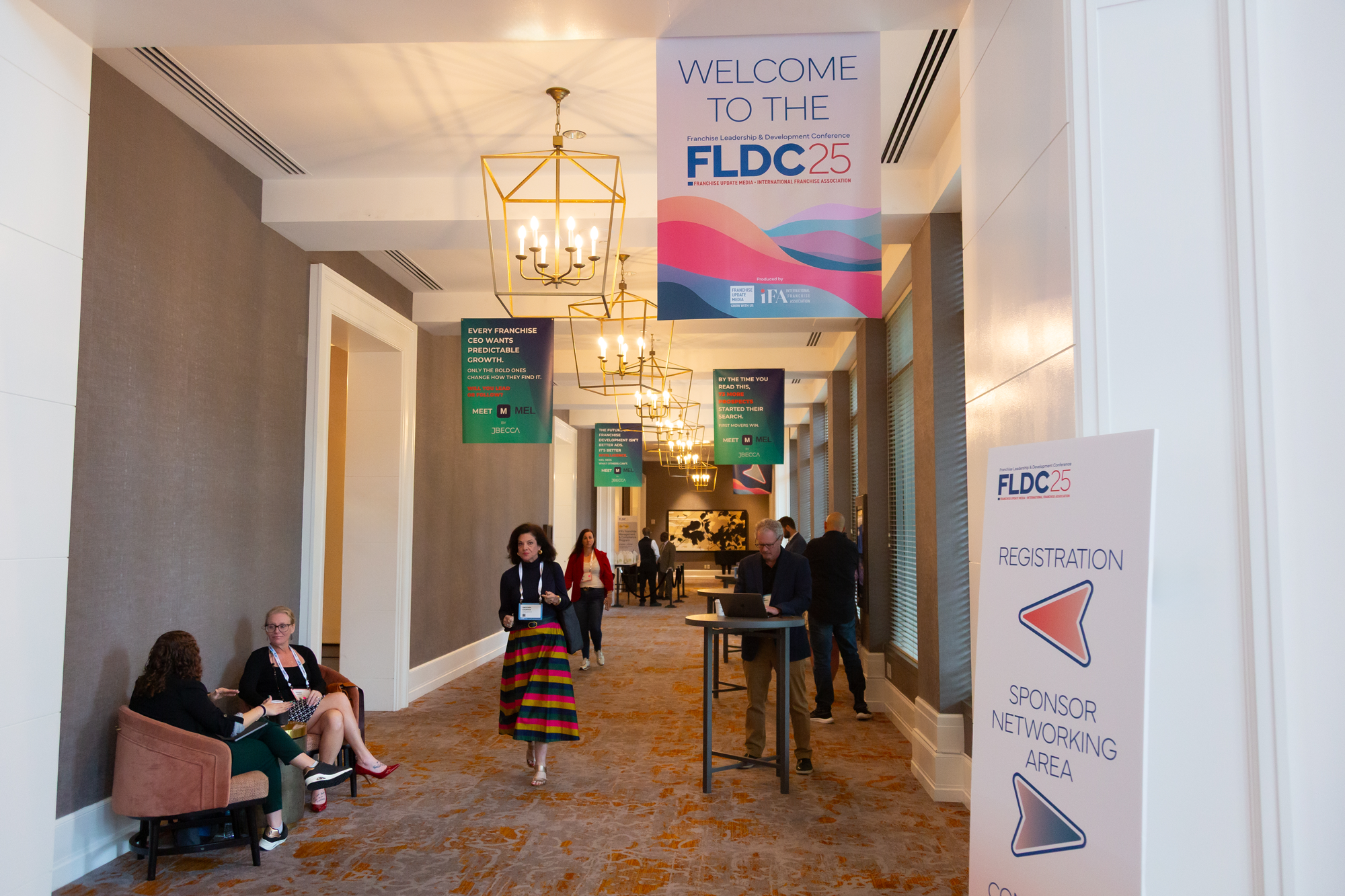 Conference hallway with people walking and sitting, large banners hanging from the ceiling reading 'Welcome to the FLDC 25' and information about event activities, with modern lighting fixtures and a carpeted floor.