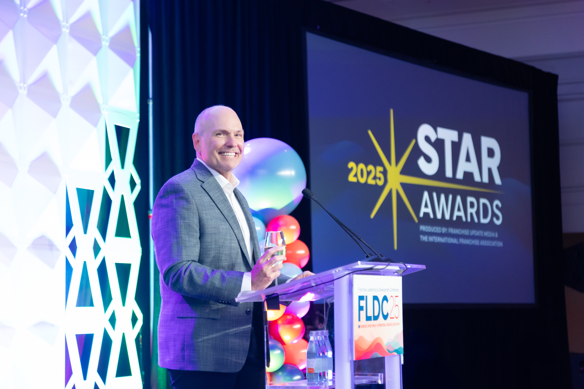 A man in a gray plaid suit holding a glass of champagne, standing at a podium during an awards event in a banquet hall. Behind him, there is a large screen displaying the event name "STAR 2025 AWARDS" and colorful balloons decorating the stage.