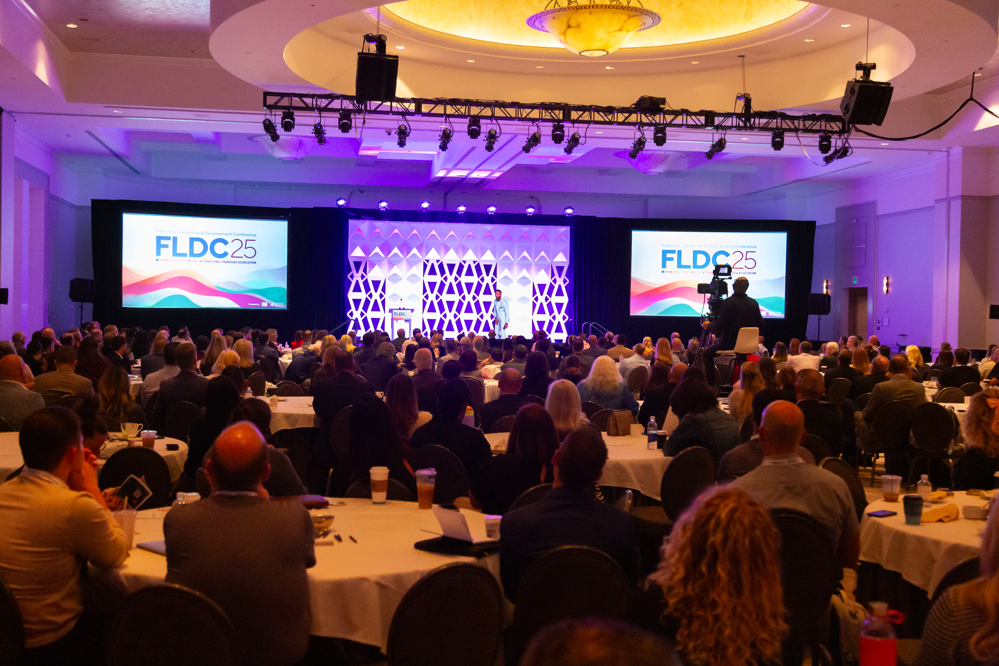 A large conference hall filled with seated attendees listening to a speaker on stage. The stage has two large screens displaying the event branding, 'FLDC 25,' with colorful wave graphics. The ceiling features a decorative circular light fixture and stage lighting. There are camera operators capturing the event.