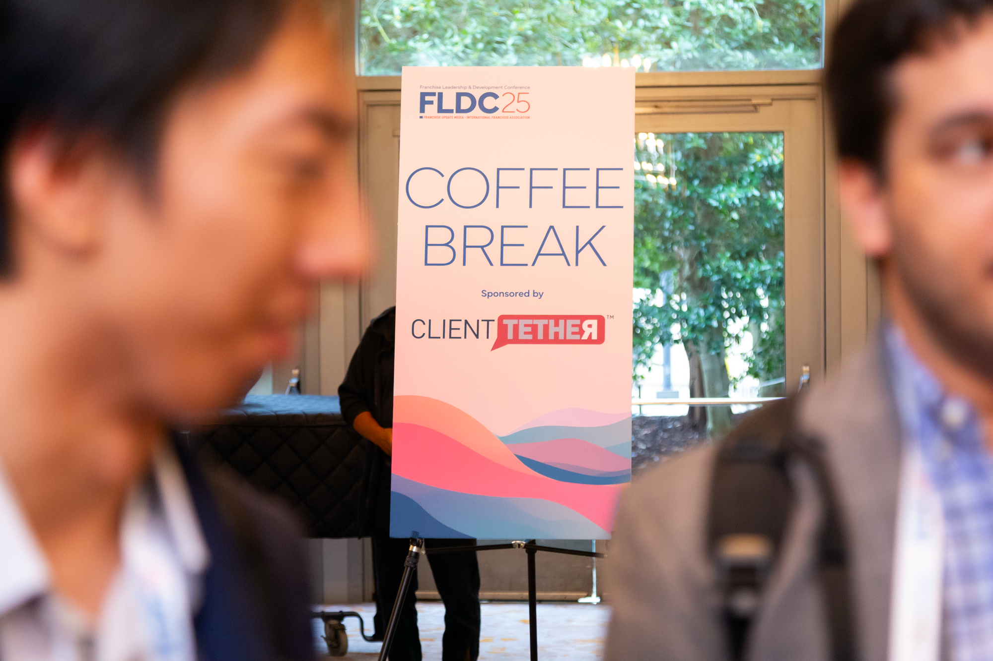 A sign on a tripod at a conference reads "Coffee Break" with event details. There are two blurred people in the foreground, with a window showing green foliage behind the sign.