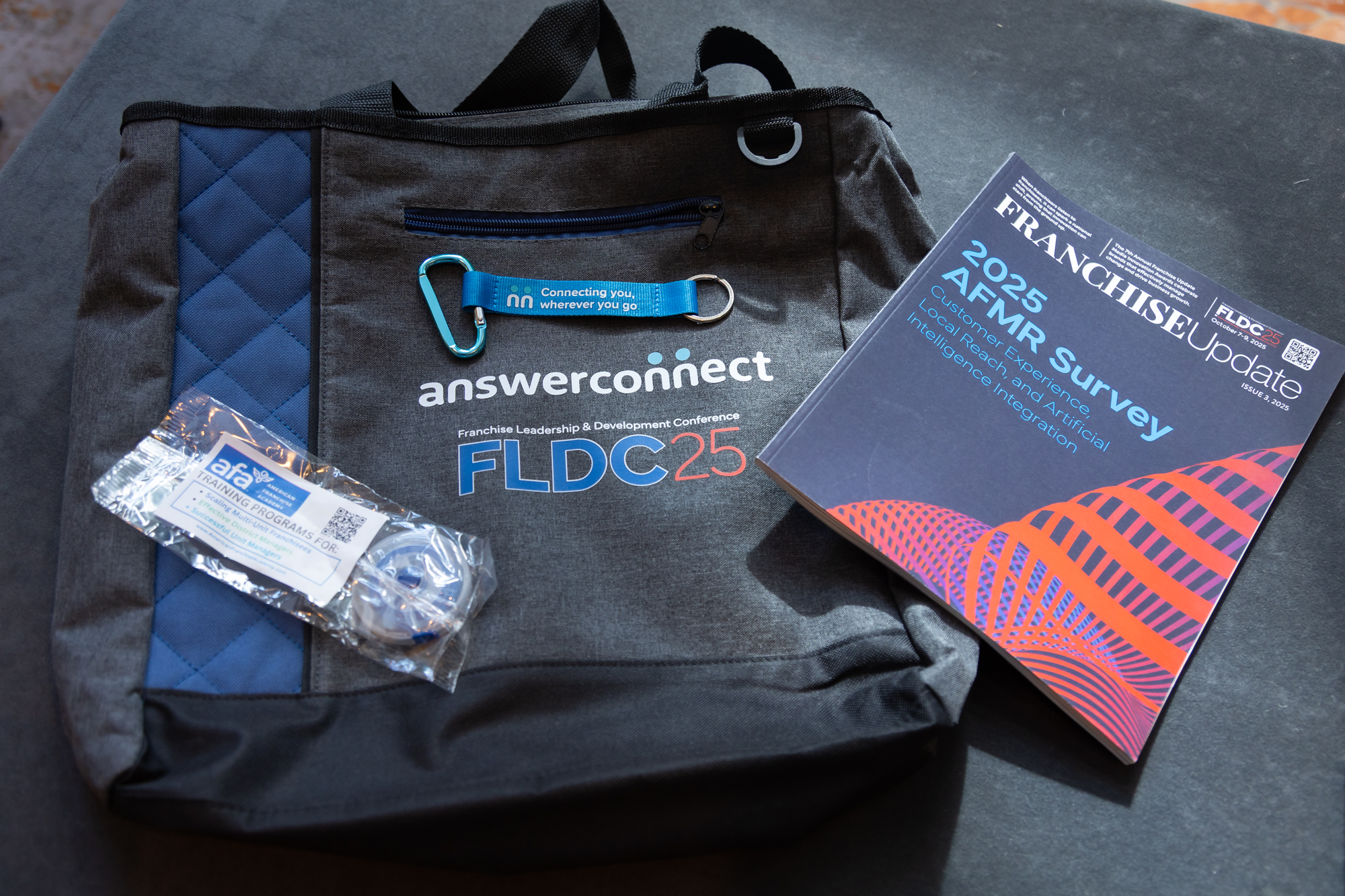 A conference bag with the AnswerConnect logo, a blue lanyard that says 'Connecting you, wherever you go,' a clear plastic badge holder, a brochure titled '2025 AFMR Surveillance,' and a small packet labeled 'AFA Training Program' placed on a dark surface.