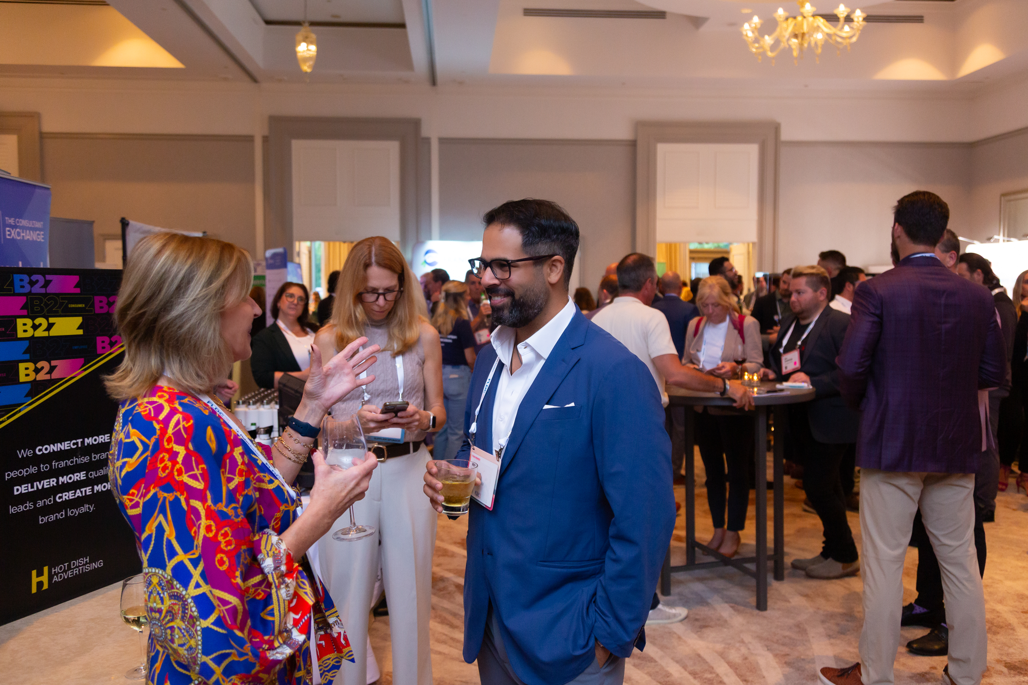 A crowded indoor networking event with people conversing, including a woman in a colorful dress and a man in a blue suit. Some are looking at their phones, and others are holding drinks. There are booths and banners in the background.