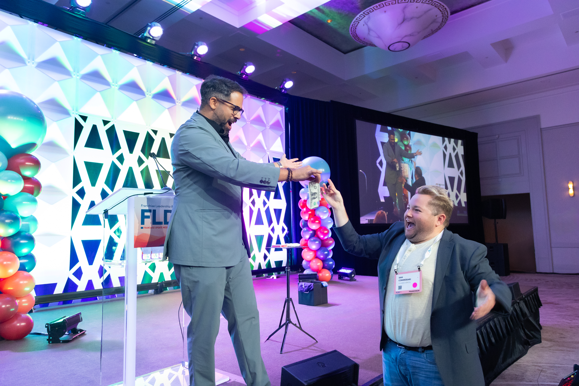 Two men at a conference or event, with one man in a gray suit handing money to the other man in a darker suit who is sitting and smiling, while balloons and a decorated backdrop are in the background.