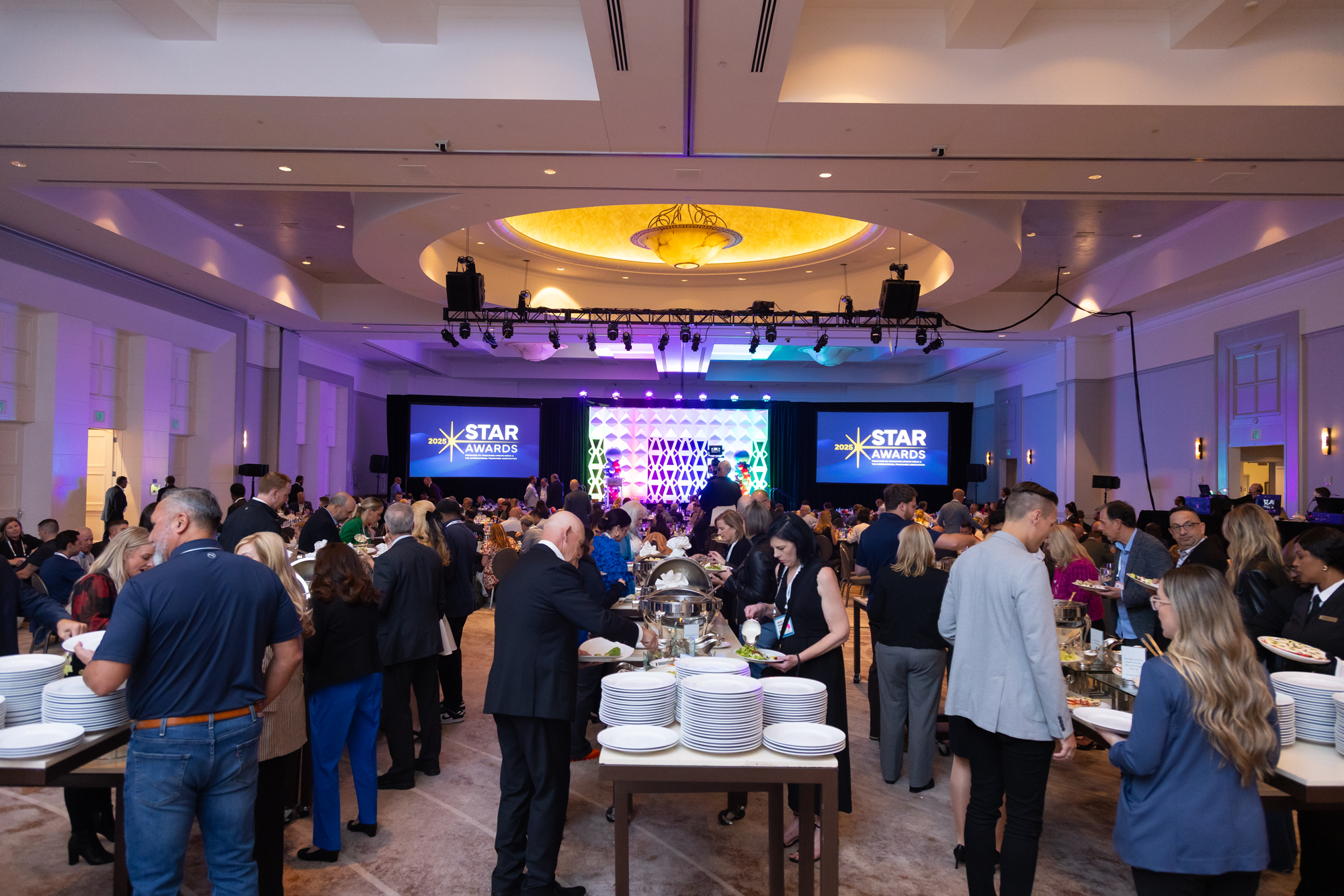 A large group of people dressed in formal attire attending a banquet in a hotel ballroom for the 2025 STAR Awards event, with tables of food, a stage with multiple large screens displaying event branding, and purple stage lighting.