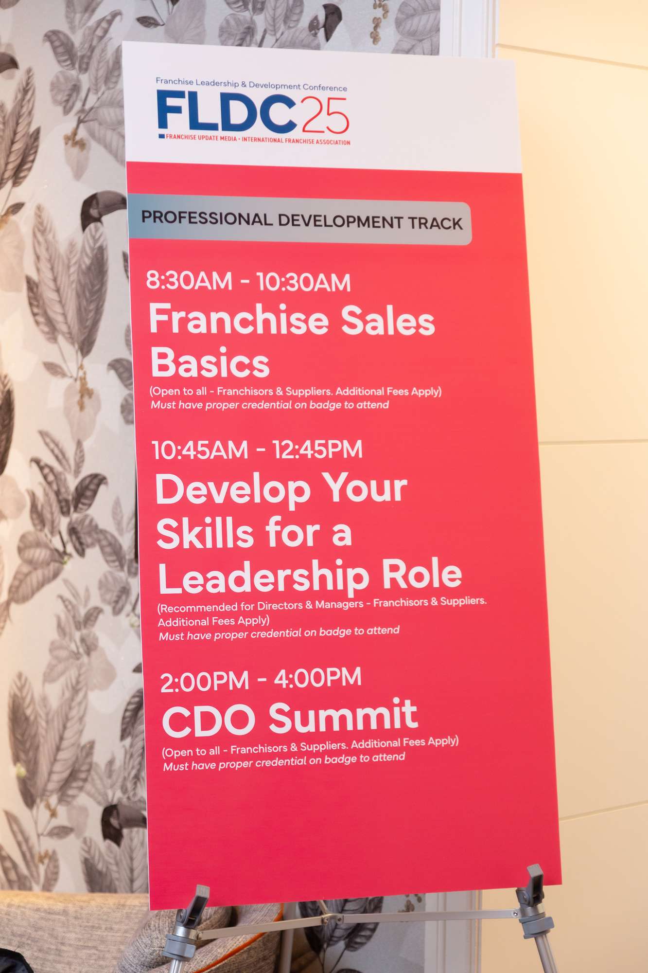 A conference schedule poster on a tripod stand. The poster has a red background with white and blue text about various sessions, including franchise sales basics, develop your skills for a leadership role, and CDO summit. The top features the logo for FDC25, the Franchise Leadership & Development Conference.