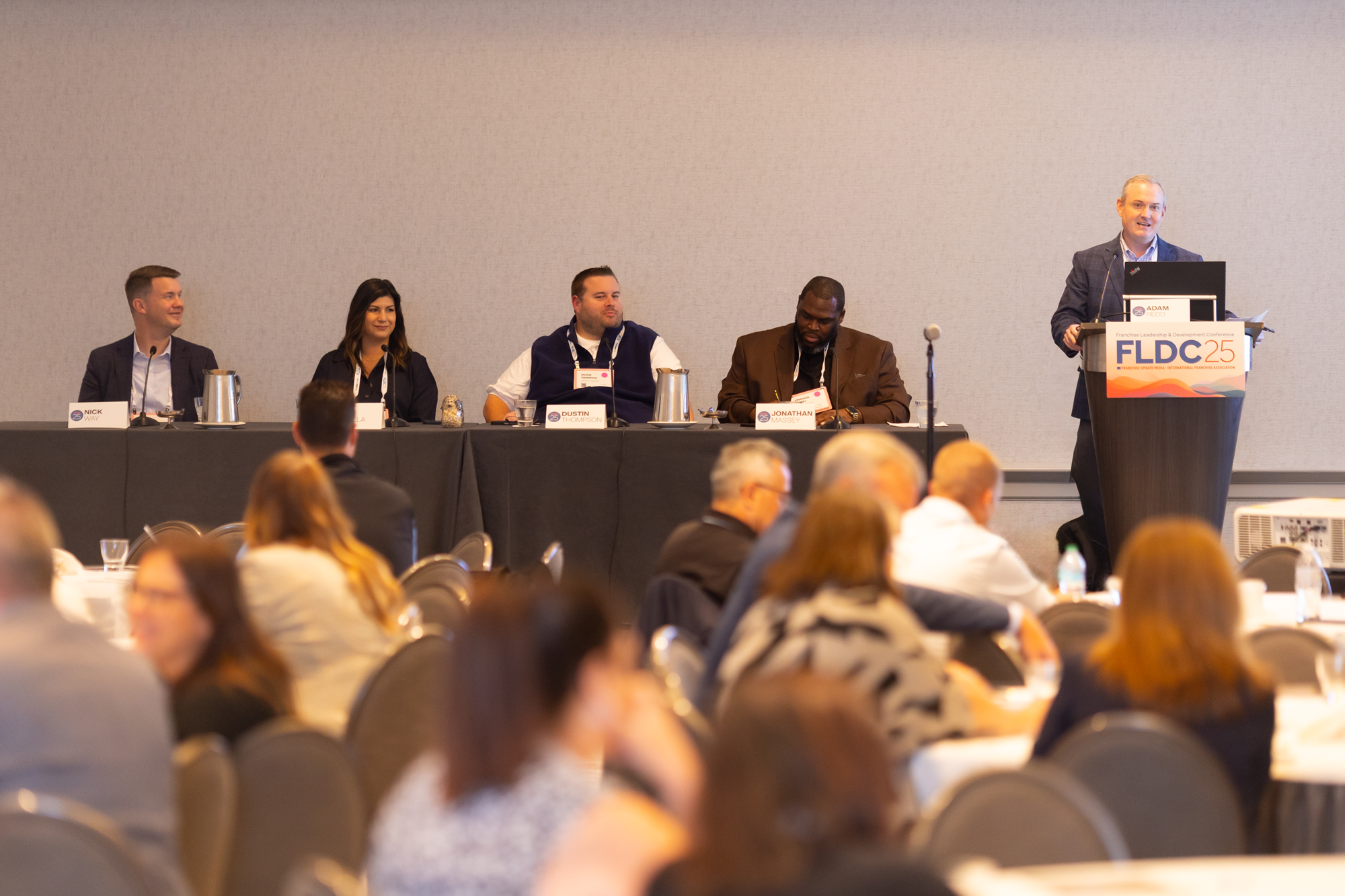 A man is speaking at a podium during a conference, with four panelists seated at a table behind him. The audience is visible in the foreground.